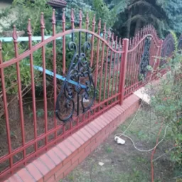 Czerwono-czarne, metalowe ogrodzenie z dekoracyjnymi elementami, zamontowane na murku z cegły w Łowiczu, z widocznymi kablami i przedłużaczem leżącymi na trawie.