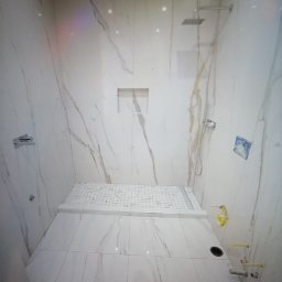 Lux-mig - Nowoczesna kabina prysznicowa wyłożona białymi płytkami imitującymi marmur, z deszczownicą i odpływem liniowym, widoczne żółte rurki instalacji wodnej.