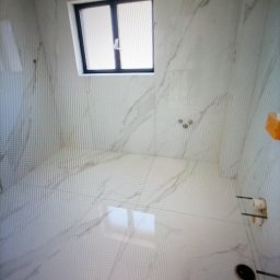 Lux-mig - Wnętrze łazienki wykończonej białymi płytkami imitującymi marmur, z oknem w czarnej ramie i zainstalowaną armaturą. Widoczne odpływy i uchwyt na papier toaletowy.