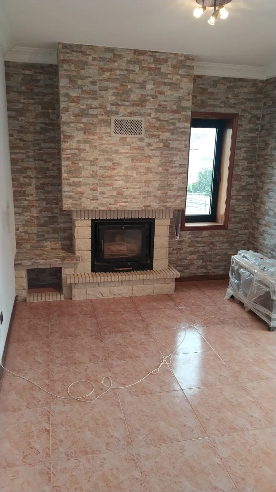 Wykończony kominek z obudową z płytek imitujących kamień, z boczną półką, w pokoju z terakotową podłogą i oknem w czarnej ramie, widok na fragment wnętrza po remoncie.