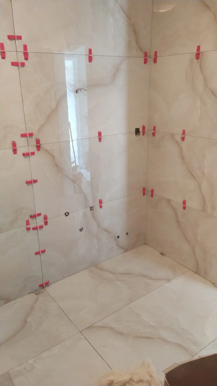 Ściany i podłoga łazienki w trakcie układania płytek imitujących marmur, z widocznymi czerwonymi klipsami poziomującymi i sznurkami, otwory na instalacje widoczne na ścianie.