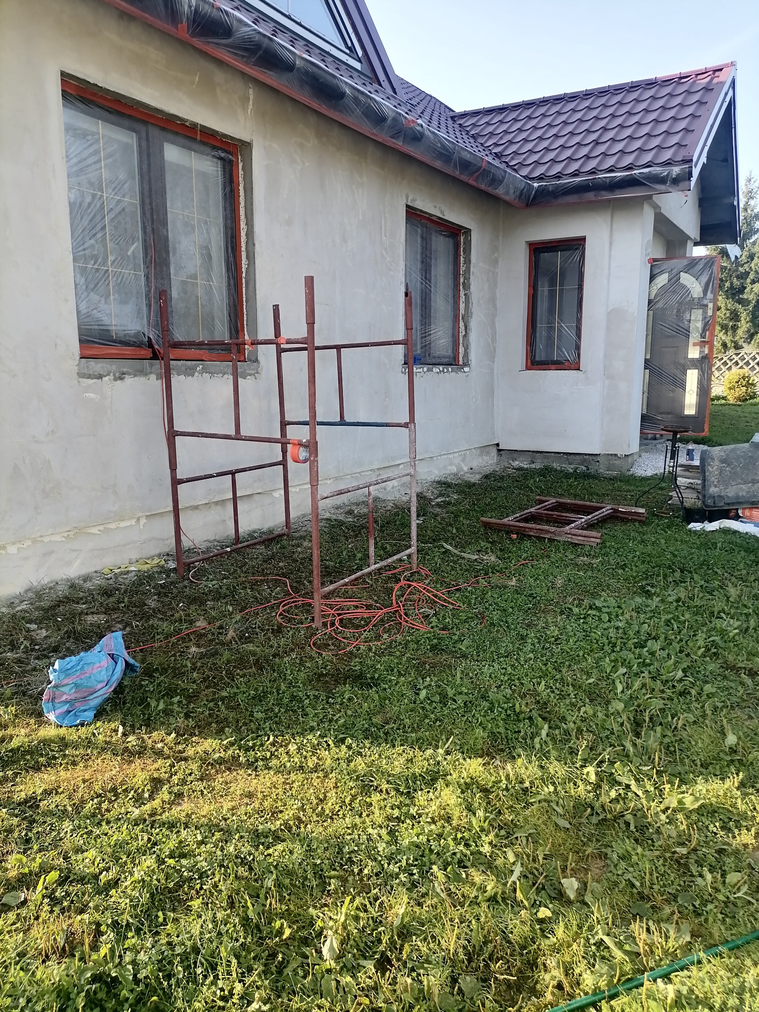 Dom jednorodzinny w trakcie prac elewacyjnych, okna zabezpieczone folią, rozstawione rusztowanie, widoczne przewody elektryczne i torba budowlana na trawie.