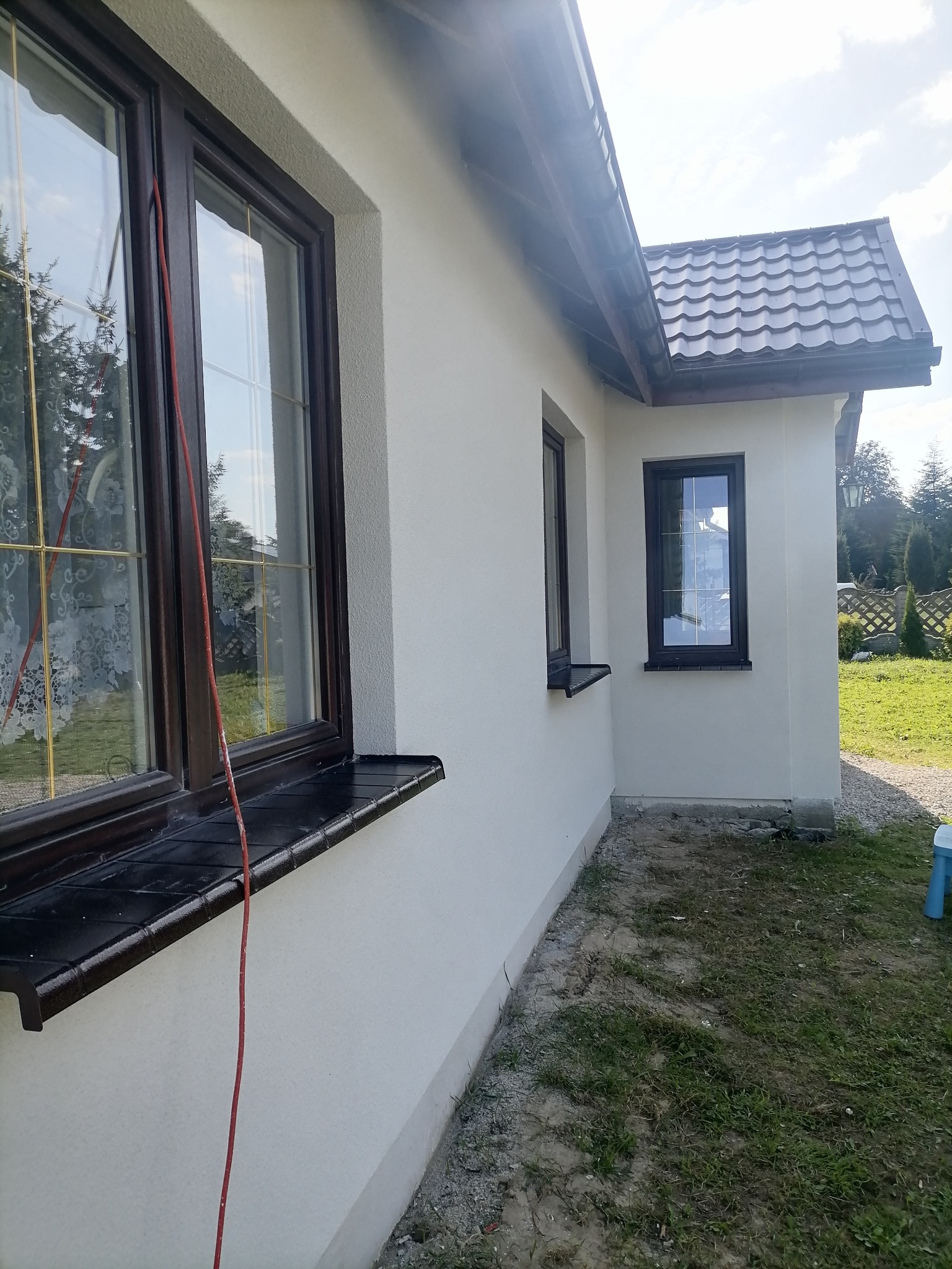 Świeżo pomalowana, jasna elewacja domu jednorodzinnego z brązowymi oknami i ciemną dachówką, widok z boku, na trawniku leży czerwony kabel.