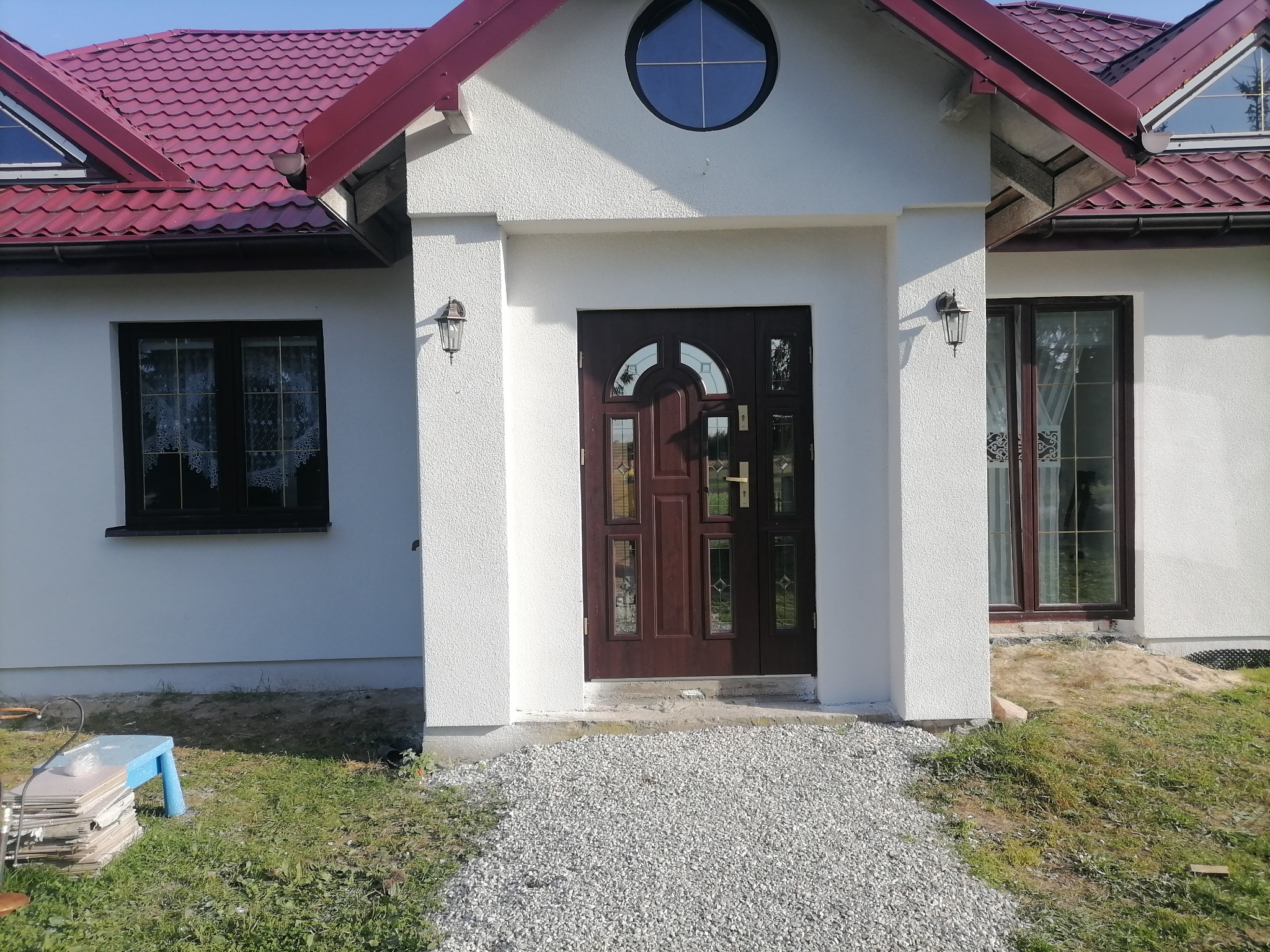 Biały dom z bordowym dachem i brązowymi drzwiami wejściowymi, okrągłe okno nad wejściem, ścieżka z białego żwiru prowadząca do drzwi.