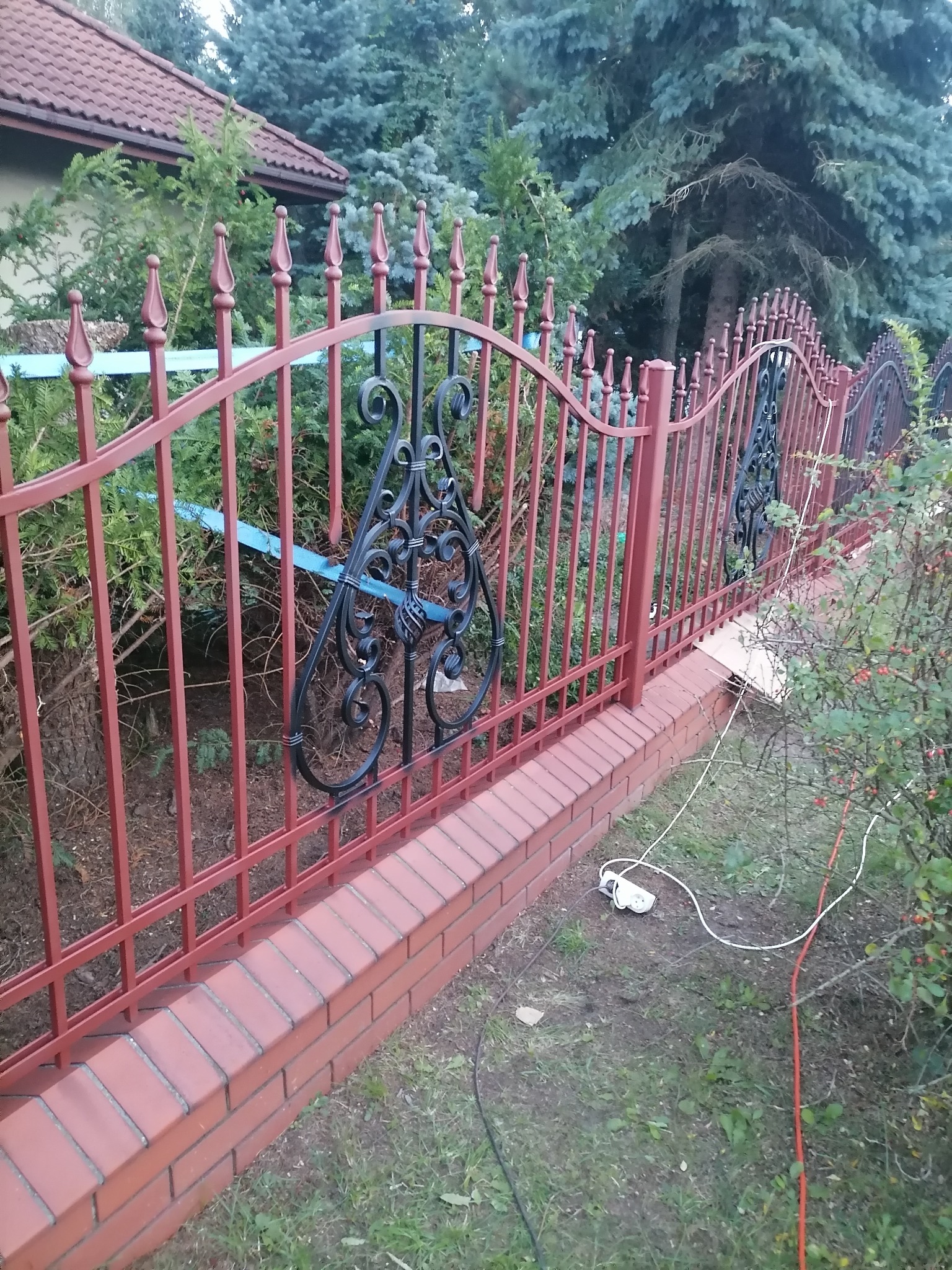 Czerwono-czarne, metalowe ogrodzenie z dekoracyjnymi elementami, zamontowane na murku z cegły w Łowiczu, z widocznymi kablami i przedłużaczem leżącymi na trawie.