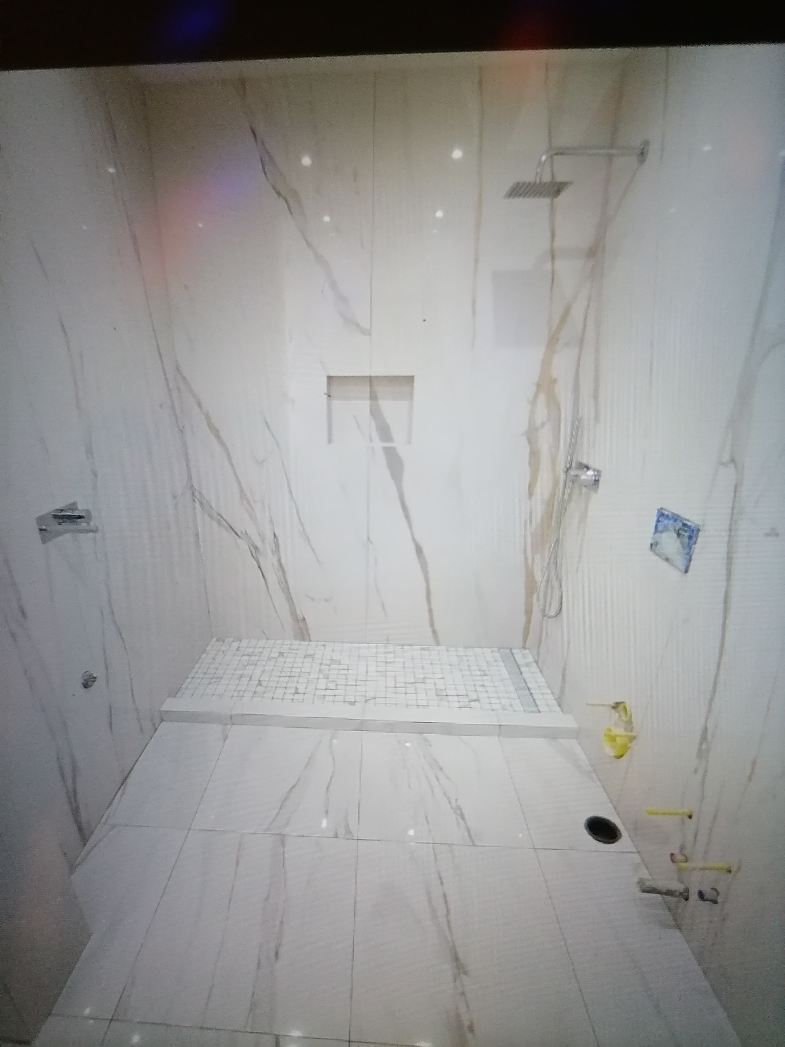 Nowoczesna kabina prysznicowa wyłożona białymi płytkami imitującymi marmur, z deszczownicą i odpływem liniowym, widoczne żółte rurki instalacji wodnej.