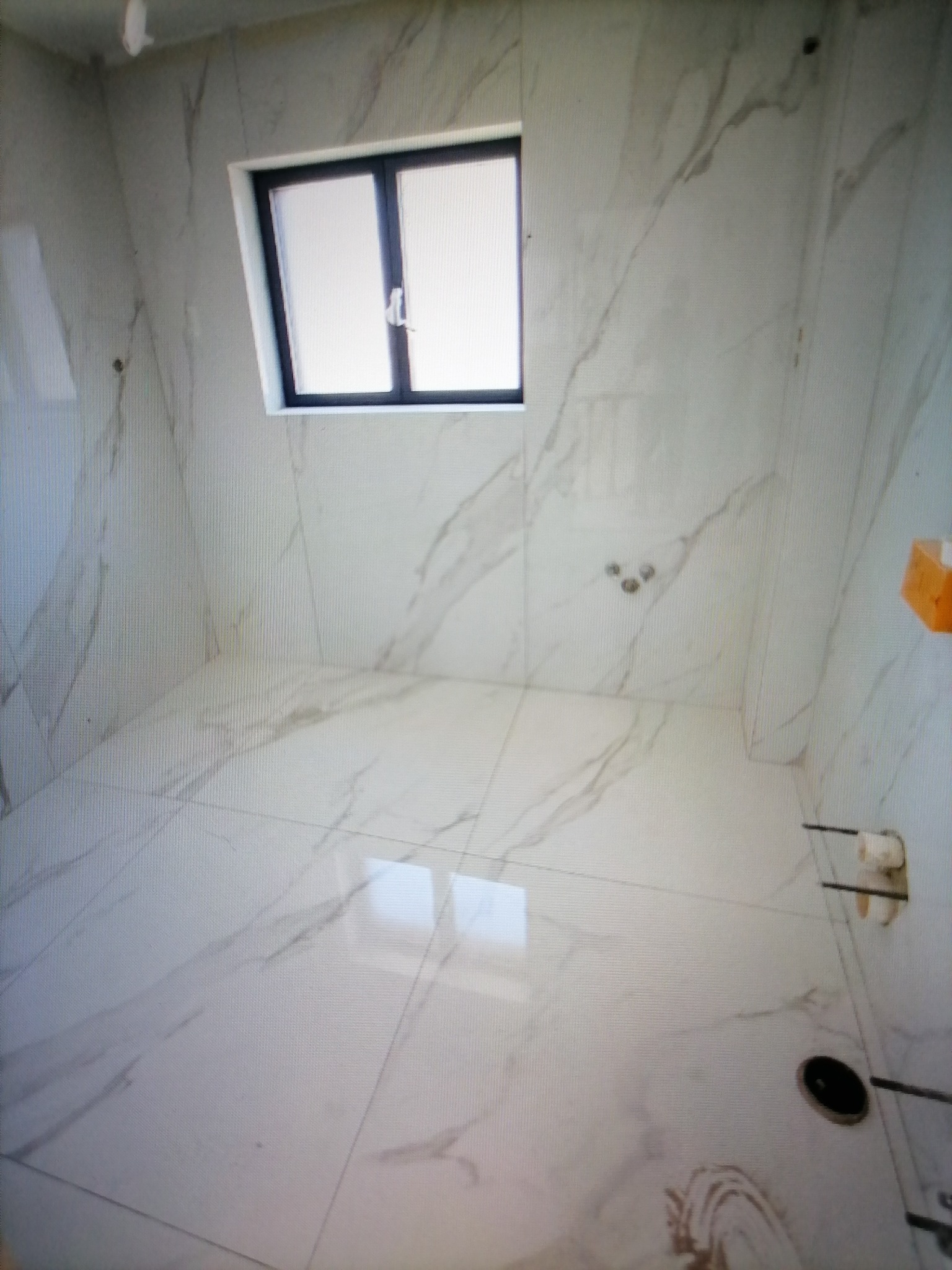 Wnętrze łazienki wykończonej białymi płytkami imitującymi marmur, z oknem w czarnej ramie i zainstalowaną armaturą. Widoczne odpływy i uchwyt na papier toaletowy.