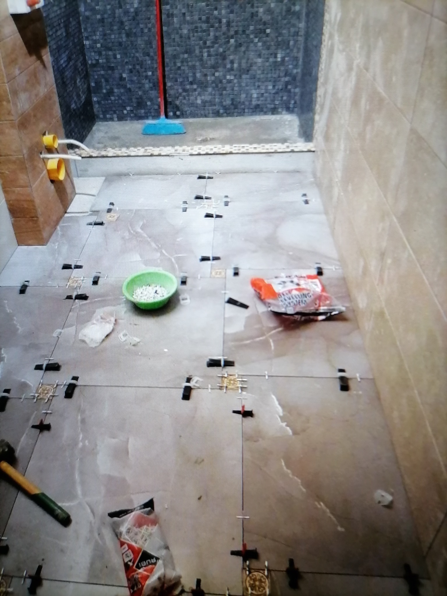 Układanie płytek podłogowych w łazience, widoczne krzyżyki dystansowe, poziomica, młotek i wiadro z zaprawą, w tle fragment ściany wyłożonej ciemnymi płytkami.