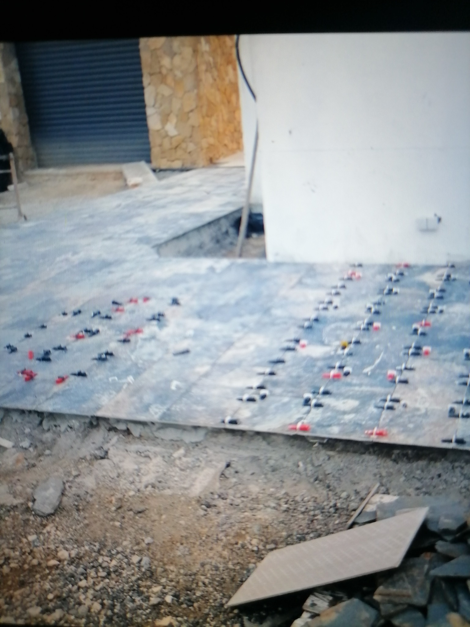 Układanie płytek na zewnątrz budynku, widoczne systemy poziomowania płytek z czerwonymi i czarnymi klinami, w tle ściana z kamienia i brama rolowana.