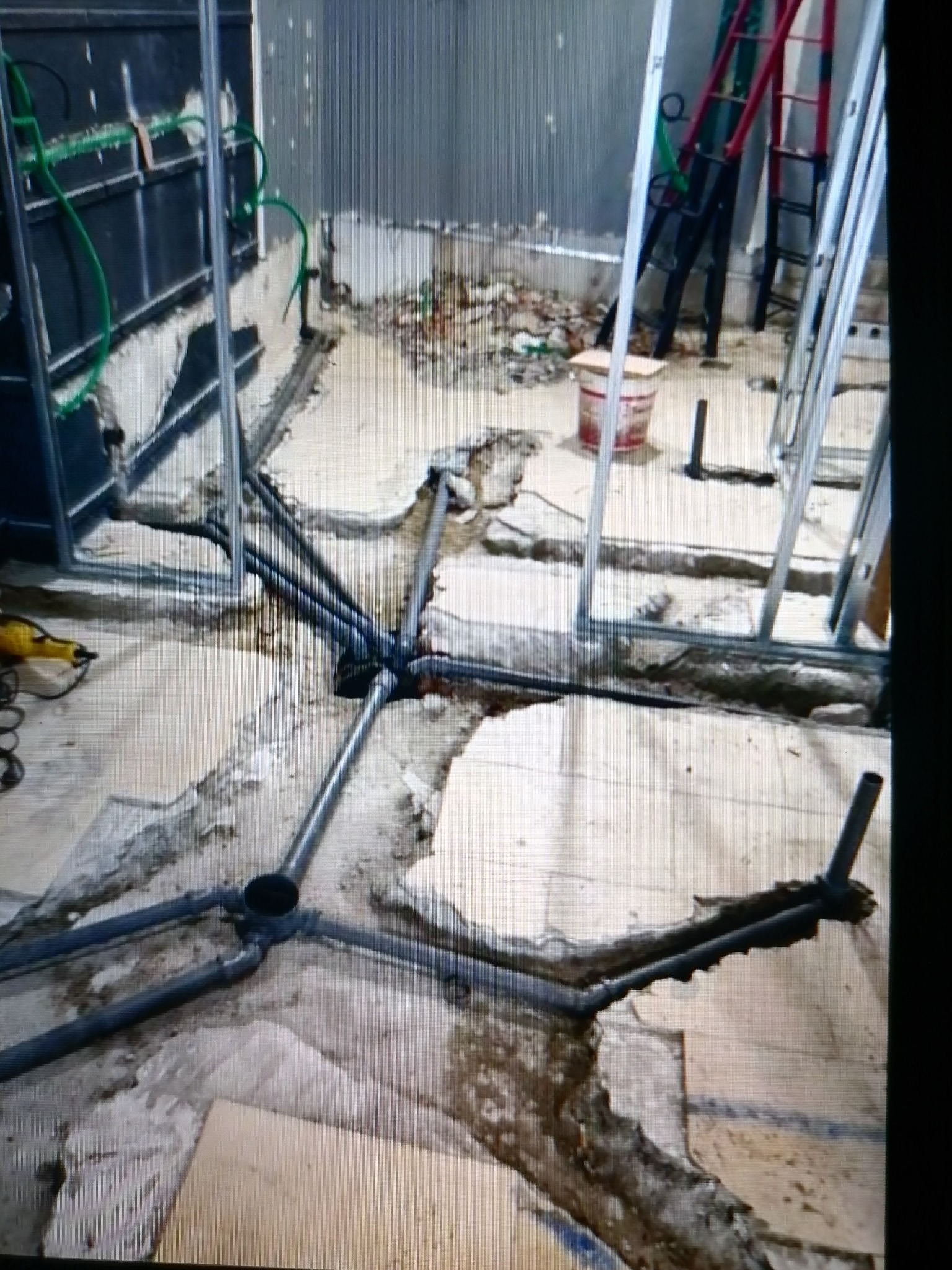 Podłoga w trakcie instalacji rur kanalizacyjnych w łazience, widoczne metalowe profile ścianek działowych, fragment drabiny i pozostałości po skuciu płytek.