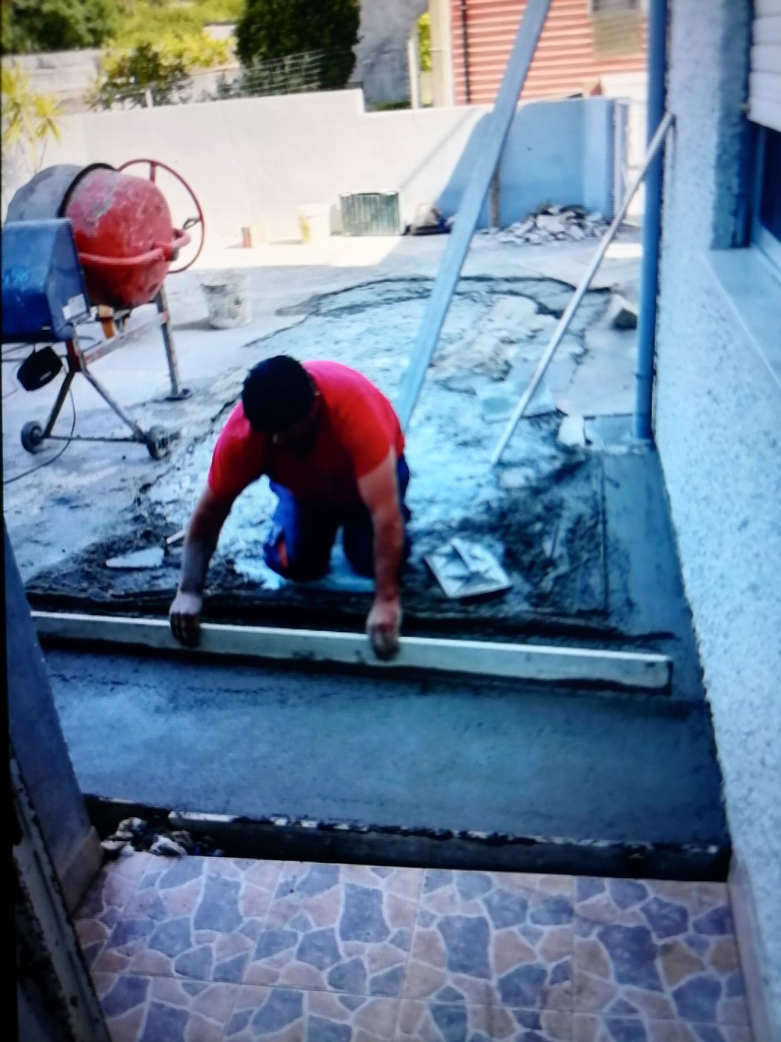 Mężczyzna w czerwonej koszulce wyrównuje wylewkę betonową przed wejściem do budynku, widoczna mieszarka do betonu i płytki ceramiczne na podłodze.