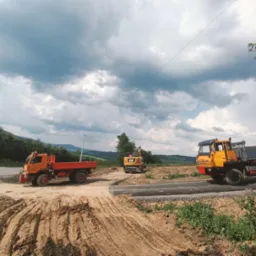 Dwa pomarańczowe samochody ciężarowe i żółta koparka CAT na placu budowy z widocznymi śladami opon w ziemi i malowniczym, pochmurnym niebem w tle.