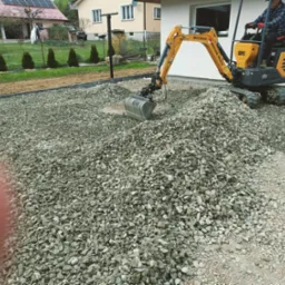 Żółta mini koparka Berger Kraus BK1250AS z operatorem w czerwonych rękawicach nabiera łyżką szarą warstwę kruszywa na posesji przy białym budynku.