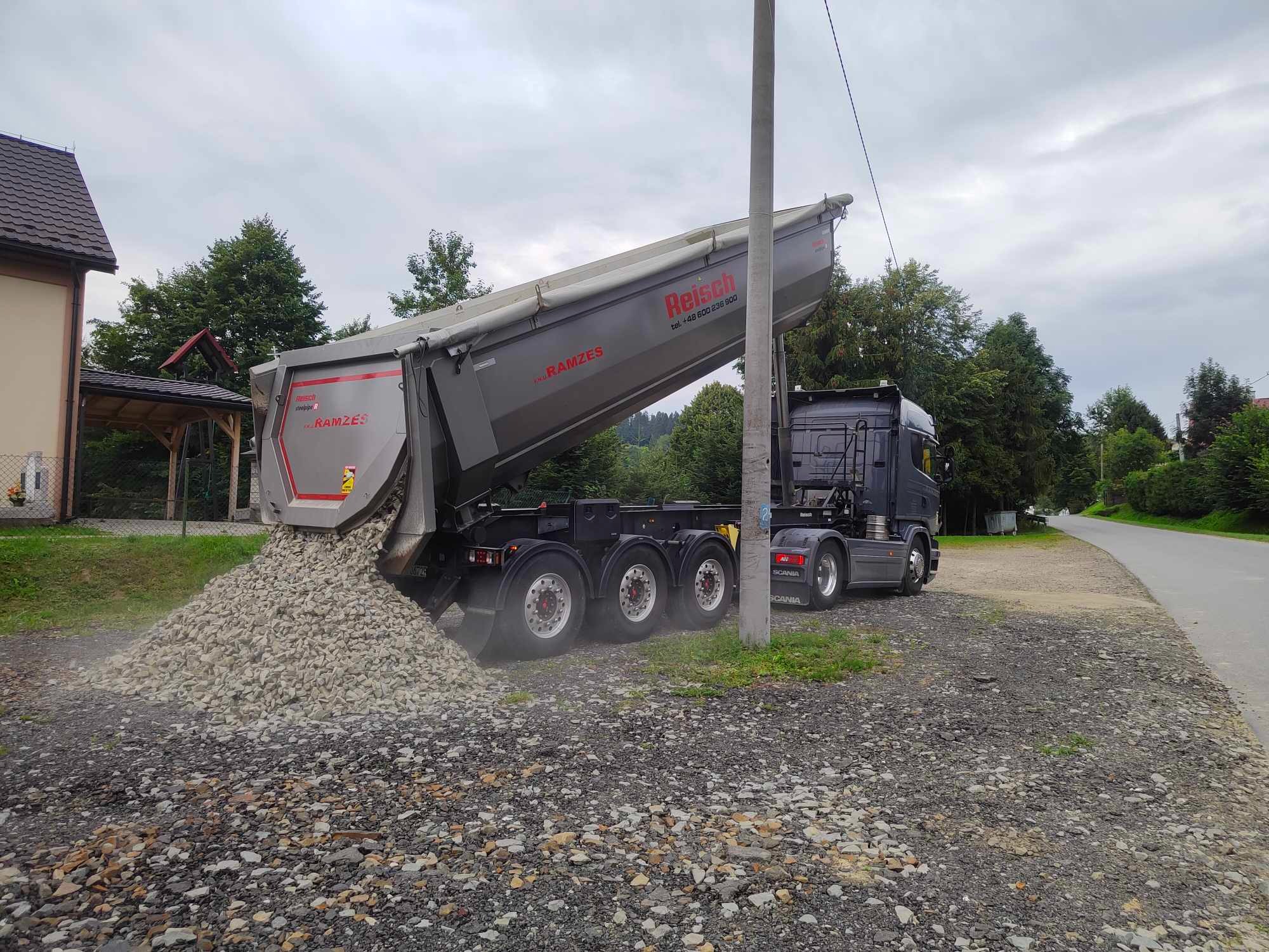 Ciężarówka wywrotka marki Scania z podniesioną skrzynią ładunkową, wysypująca ładunek żwiru na pobocze drogi w otoczeniu drzew i zabudowań.