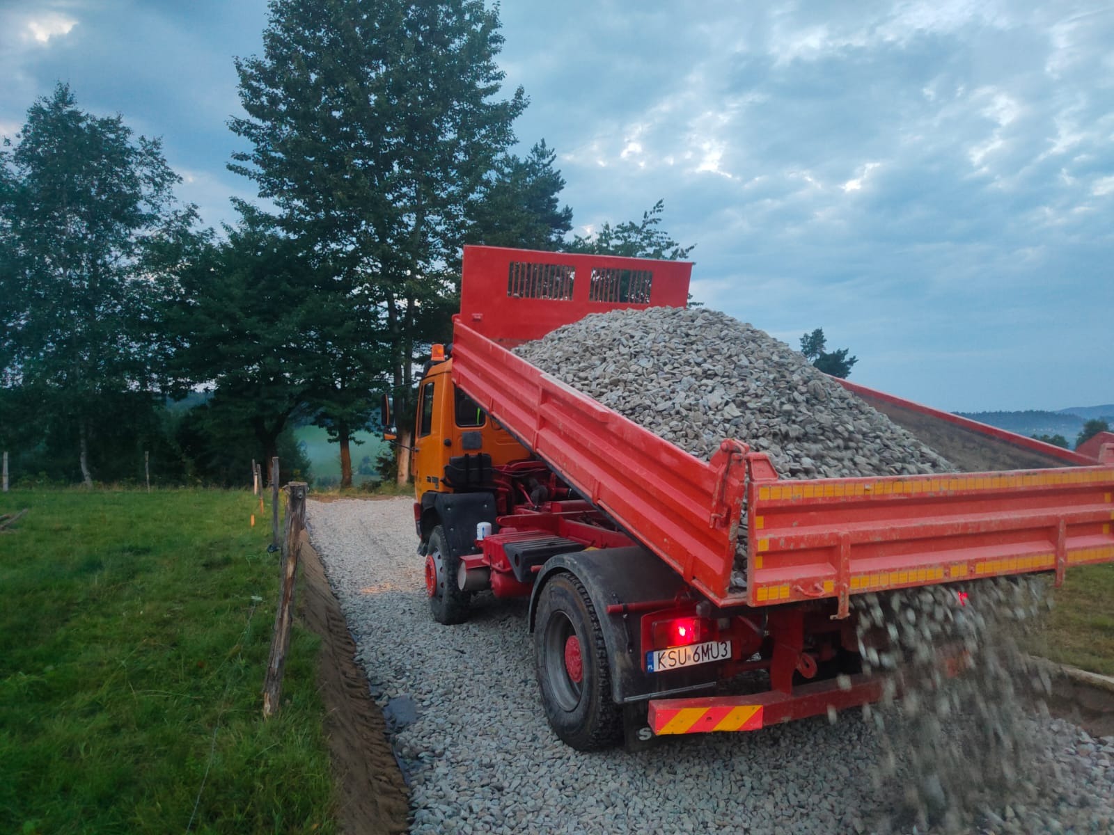 Pomarańczowo-czerwona ciężarówka wywrotka z otwartą skrzynią ładunkową, wysypująca żwir na utwardzoną drogę gruntową z widoczną na pierwszym planie świeżo przygotowaną krawędzią drogi, w tle zieleń...