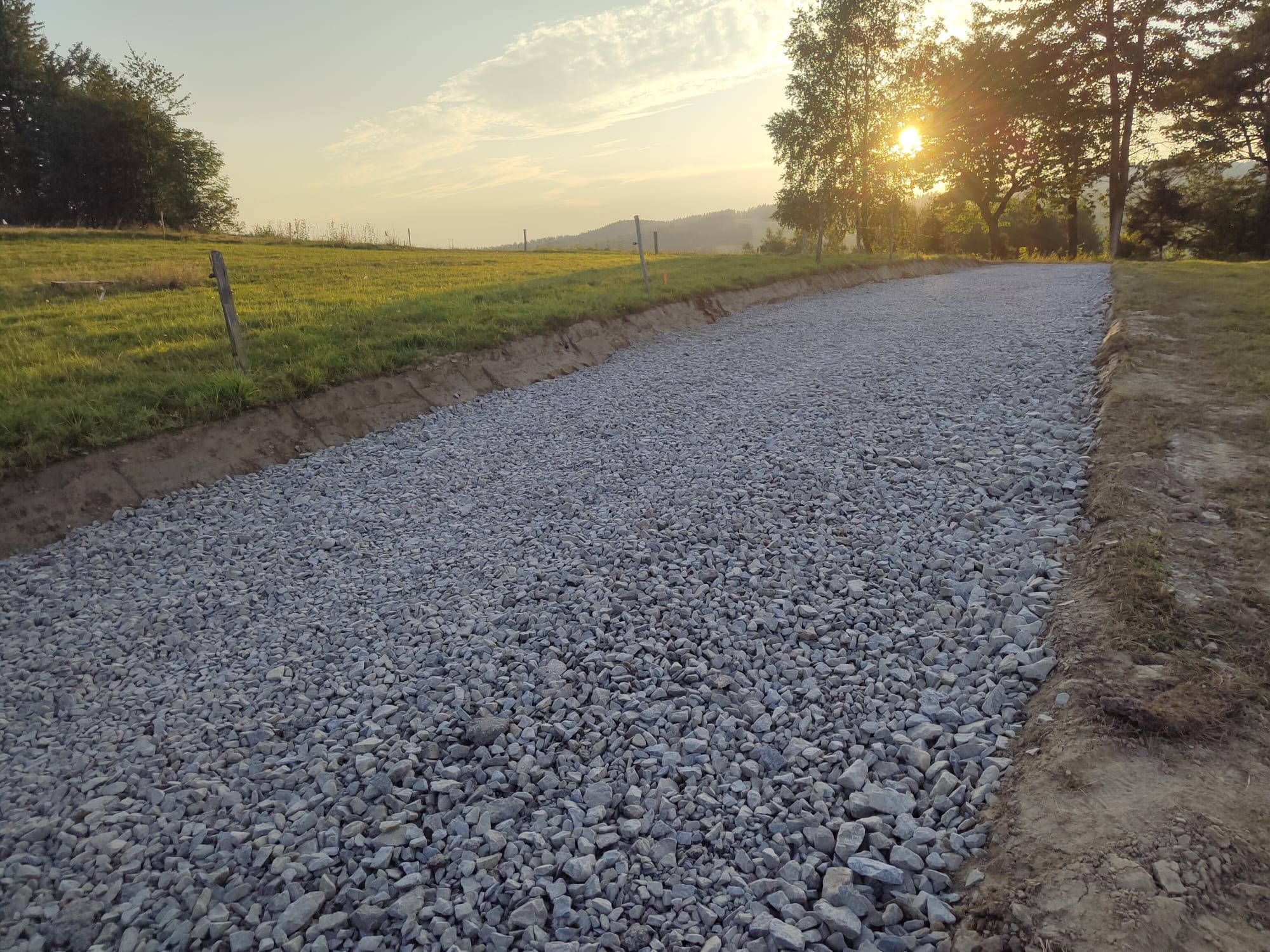 Przygotowany teren pod budowę z wysypanym kruszywem, widok na zachodzące słońce i pagórkowaty krajobraz w tle.