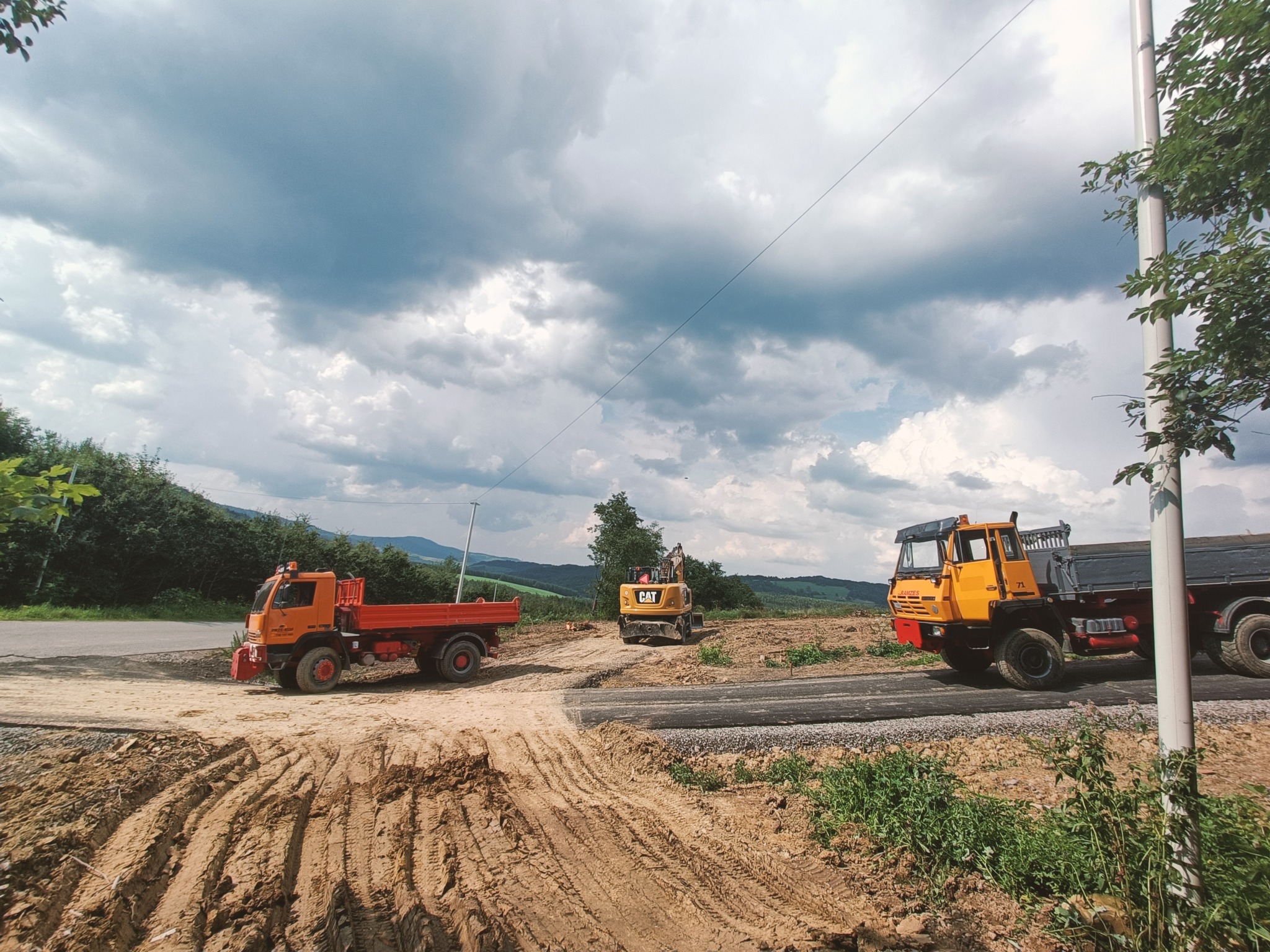 Dwa pomarańczowe samochody ciężarowe i żółta koparka CAT na placu budowy z widocznymi śladami opon w ziemi i malowniczym, pochmurnym niebem w tle.
