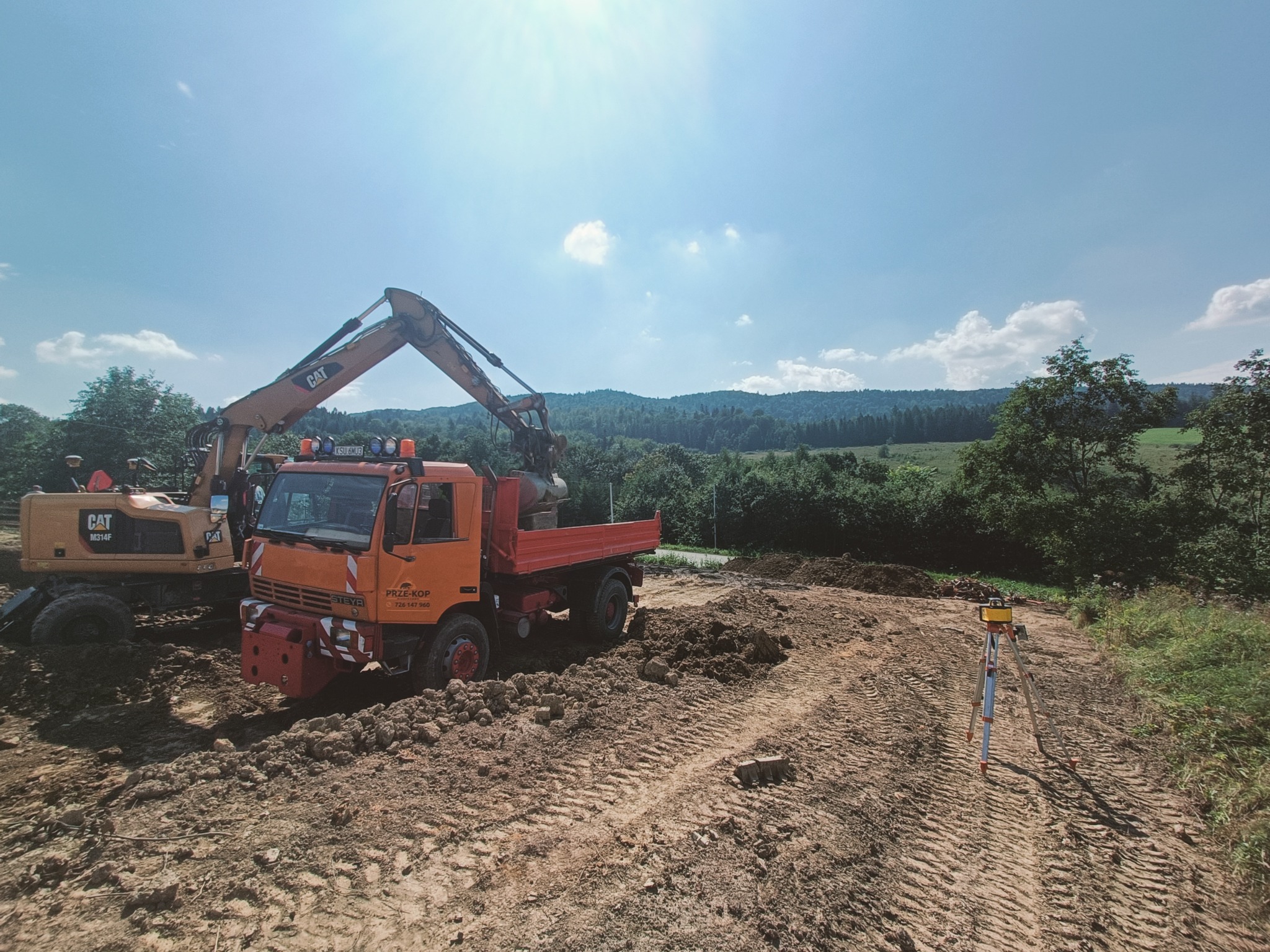 Żółty koparko-ładowarka CAT M314F i pomarańczowa wywrotka STEYR na placu budowy, z widocznym niwelatorem na statywie, w tle zalesione wzgórza i błękitne niebo z chmurami.
