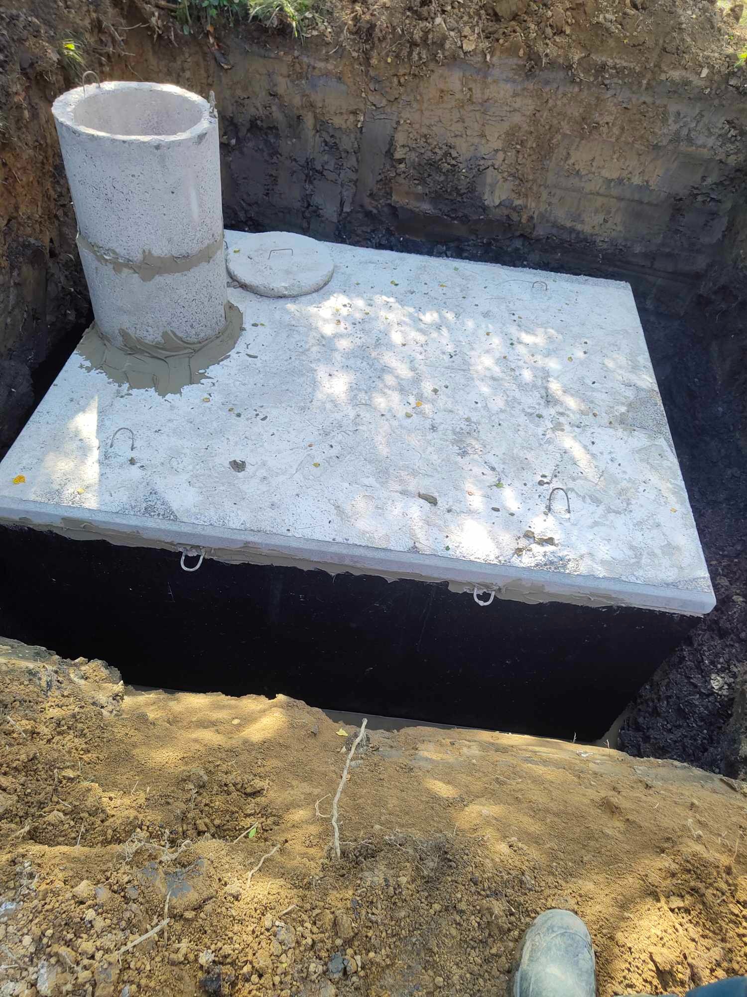 Betonowy zbiornik, prawdopodobnie szambo, umieszczony w wykopie ziemnym, z widoczną rurą wznoszącą się pionowo i pokrywą. Wokół zbiornika widoczna ziemia i fragment buta w rogu kadru.