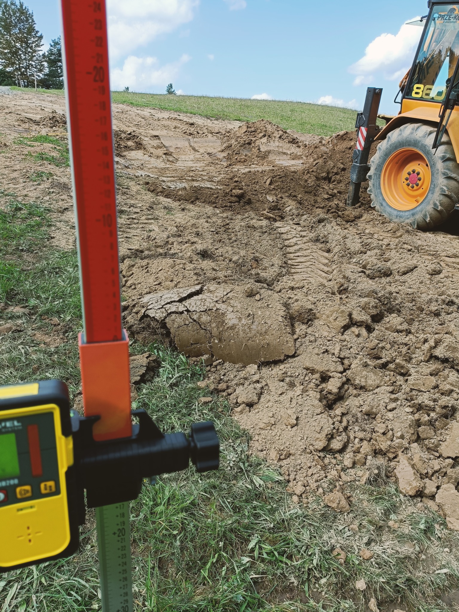 Żółta niwelator laserowy na łacie niwelacyjnej ustawiony na ziemi z wykopem w tle i częściowo widoczną koparką z tyłu, podczas prac ziemnych.