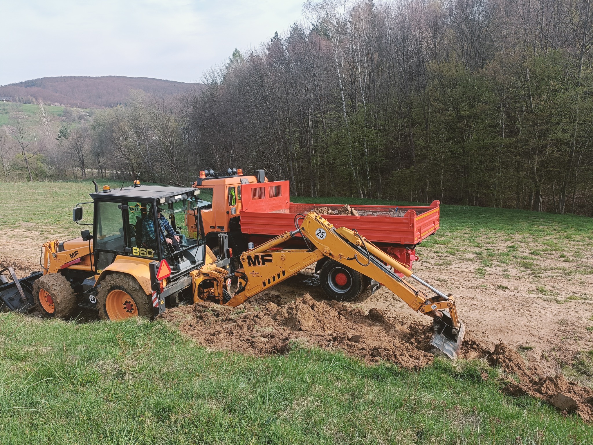 Żółto-czarna koparko-ładowarka MF 860 z operatorem, obok czerwona wywrotka, praca w terenie trawiastym z widocznymi śladami wykopu, las w tle.