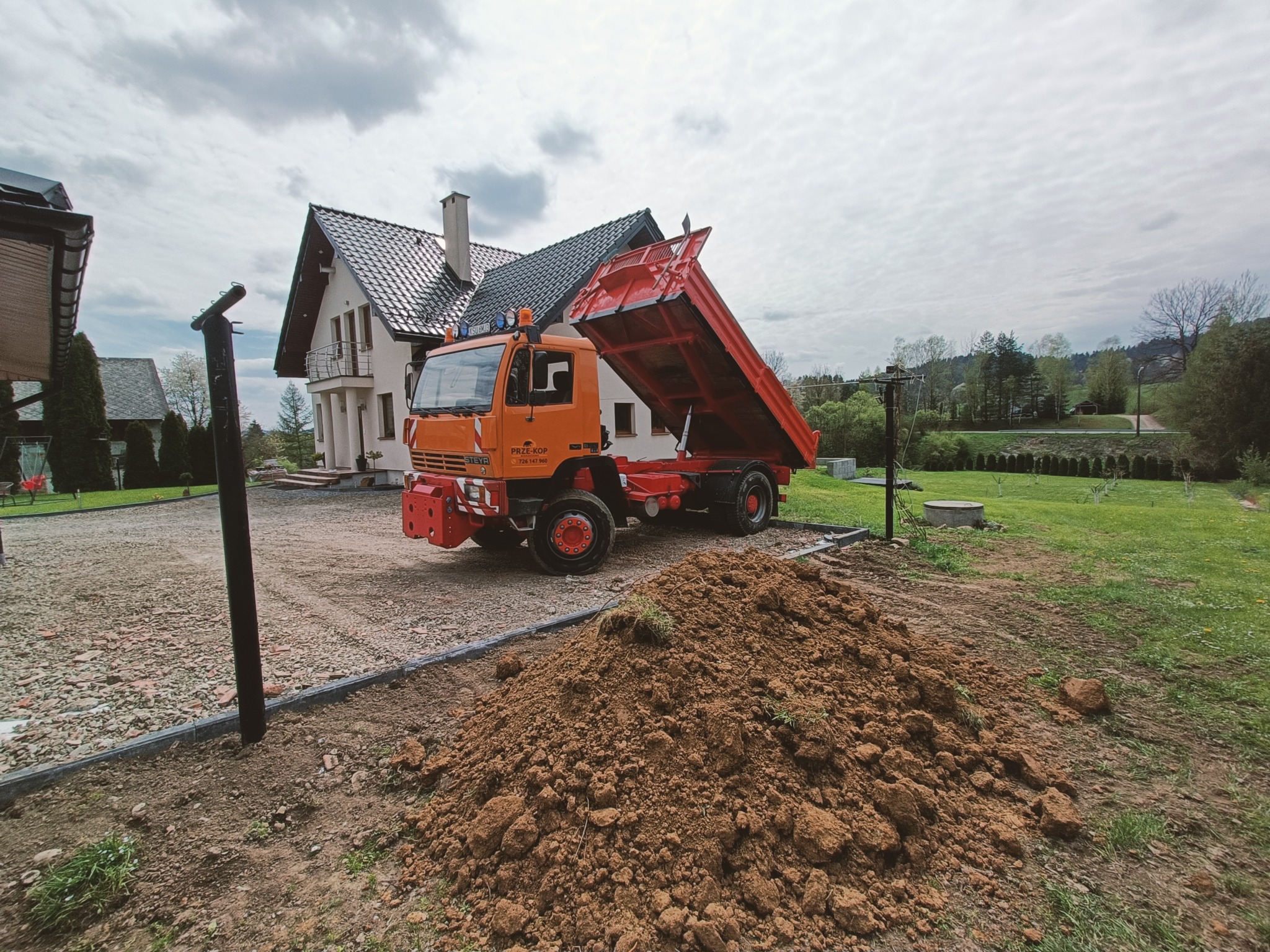 Pomarańczowy wywrotka z podniesioną skrzynią stoi obok pryzmy ziemi na tle domu jednorodzinnego i zielonego trawnika.
