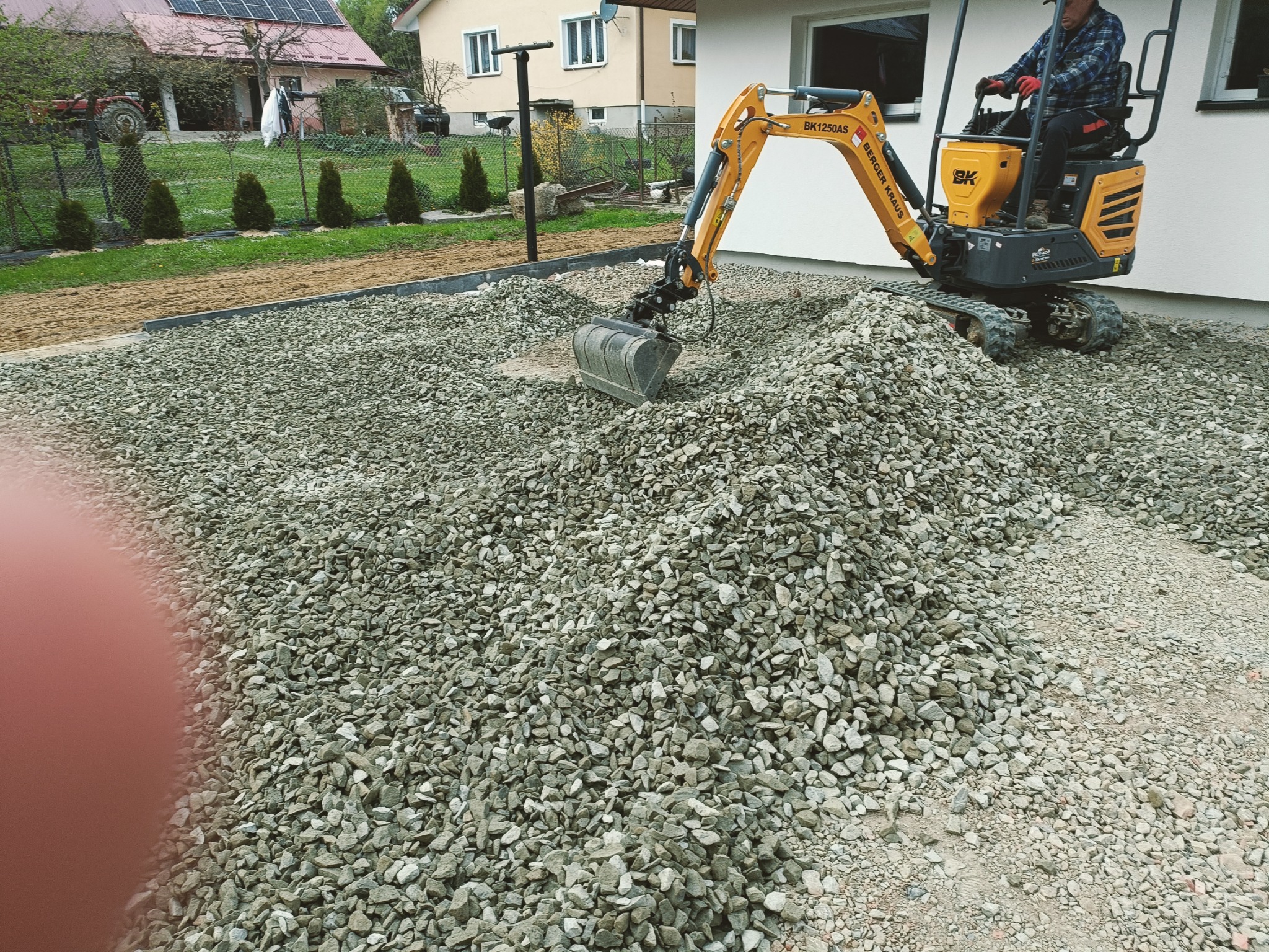 Żółta mini koparka Berger Kraus BK1250AS z operatorem w czerwonych rękawicach nabiera łyżką szarą warstwę kruszywa na posesji przy białym budynku.
