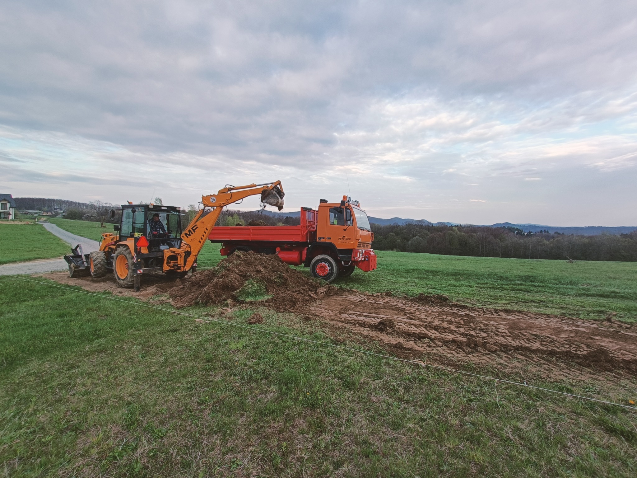Pomarańczowa koparko-ładowarka z operatorem nabiera ziemię i wysypuje ją na pomarańczową wywrotkę na tle zielonego pola i pochmurnego nieba.