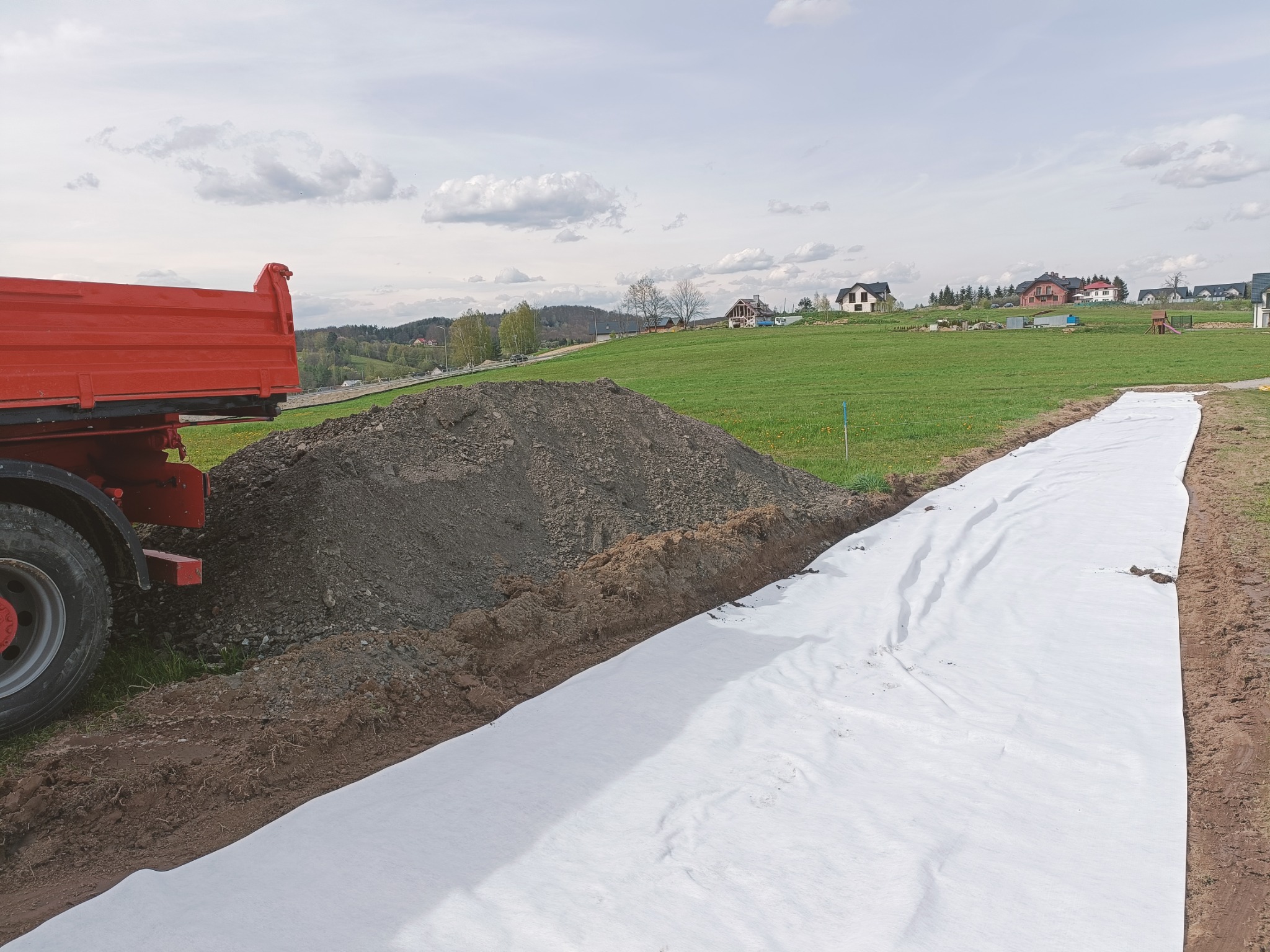 Czerwona wywrotka obok pryzmy ziemi i rozłożonej białej geowłókniny, przygotowanie podłoża na działce z widokiem na domy w oddali.