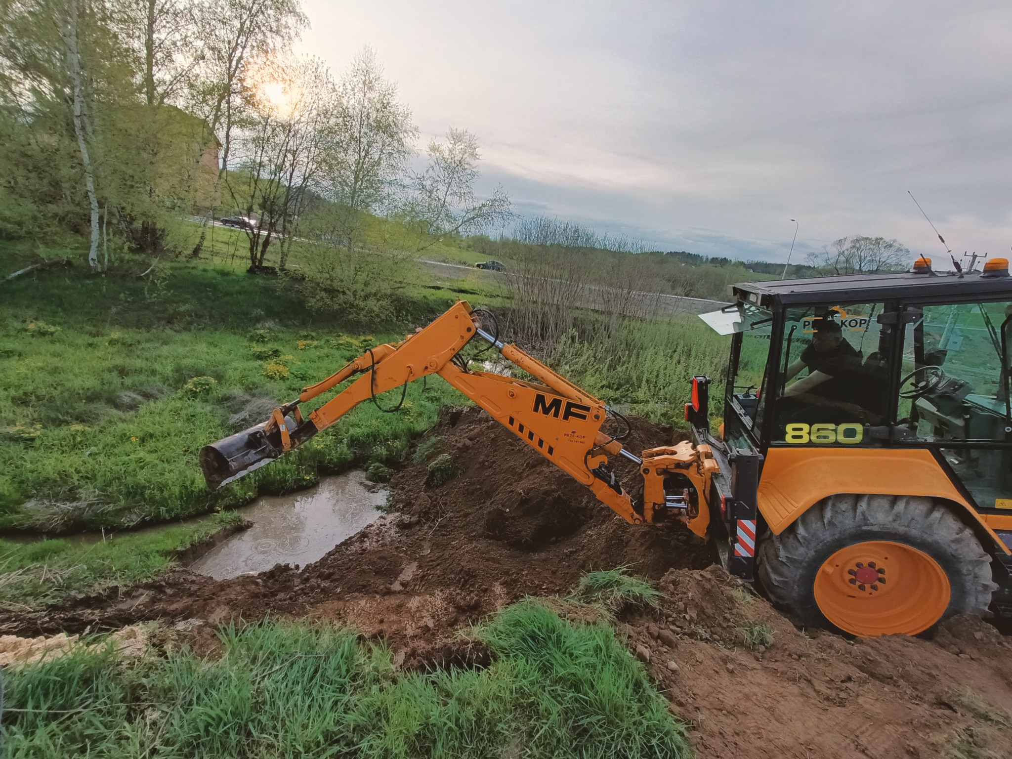 Pomarańczowa koparko-ładowarka MF 860 przy wykopie w pobliżu rowu z wodą, zielona trawa, drzewa w tle, pochmurne niebo.