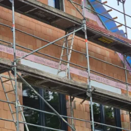 Montaż okien w surowym budynku z cegły, widoczne rusztowanie i ocieplenie w górnej części, okna z czarnymi ramami.