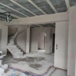 Wnętrze w stanie surowym po nałożeniu tynków: widoczne ściany i sufit pokryte tynkiem, instalacje elektryczne na suficie i podłodze, schody betonowe w tle.