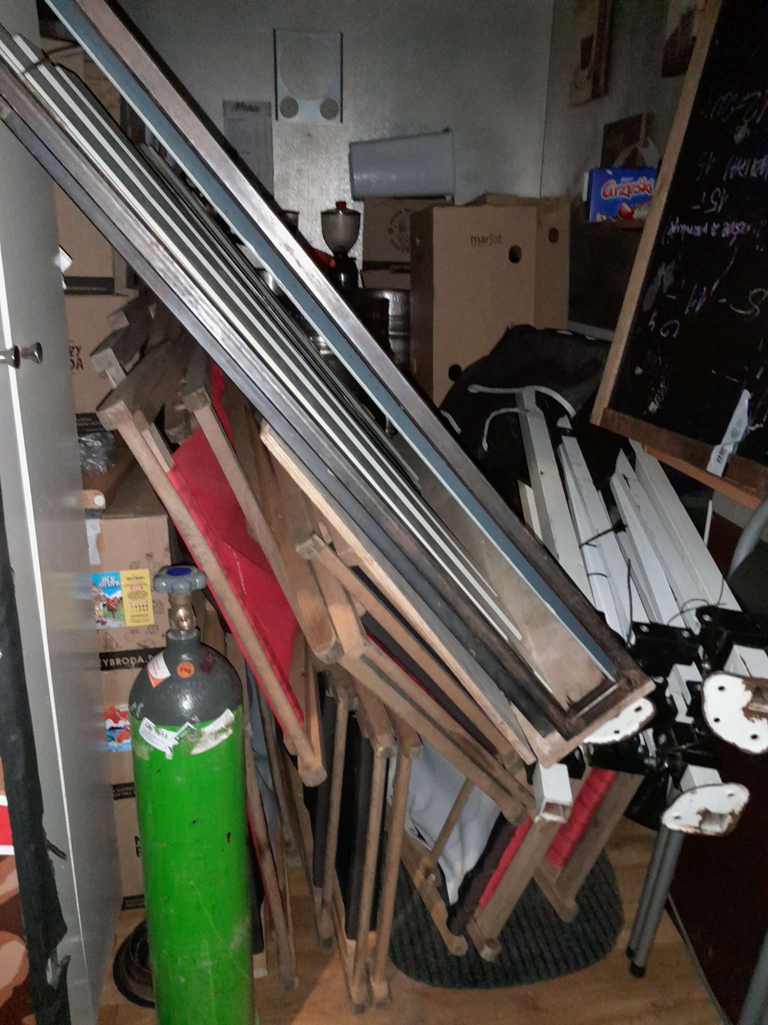 Stos złożonych, drewnianych krzeseł z czerwonymi siedziskami, opartych o ścianę, z butlą gazową w tle, w zagraconym pomieszczeniu.