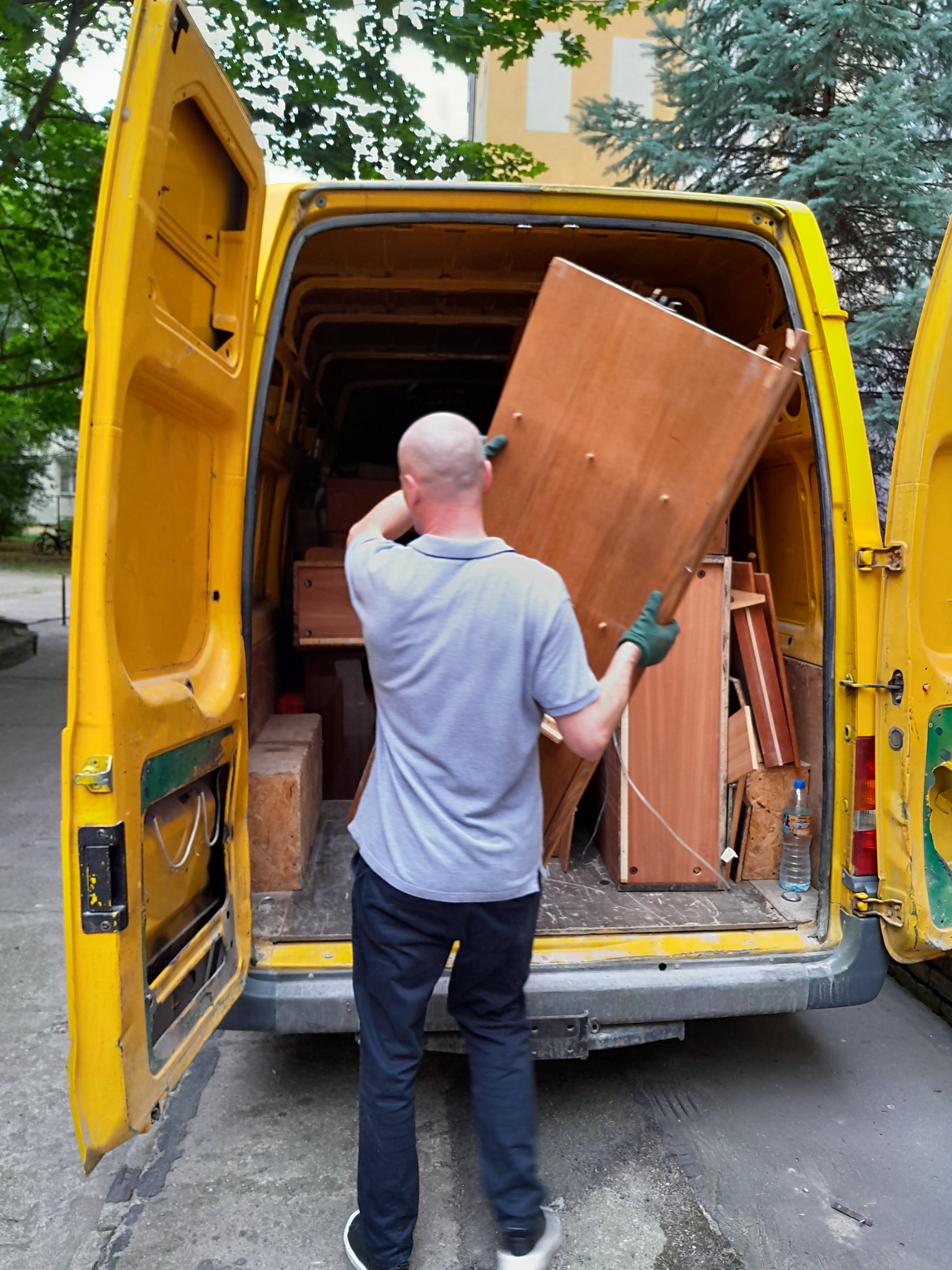 Mężczyzna w szarym T-shircie i ciemnych spodniach, w zielonych rękawicach, wnosi element mebla do żółtego busa z otwartymi drzwiami z tyłu; widoczne inne elementy mebli i butelka wody wewnątrz busa.