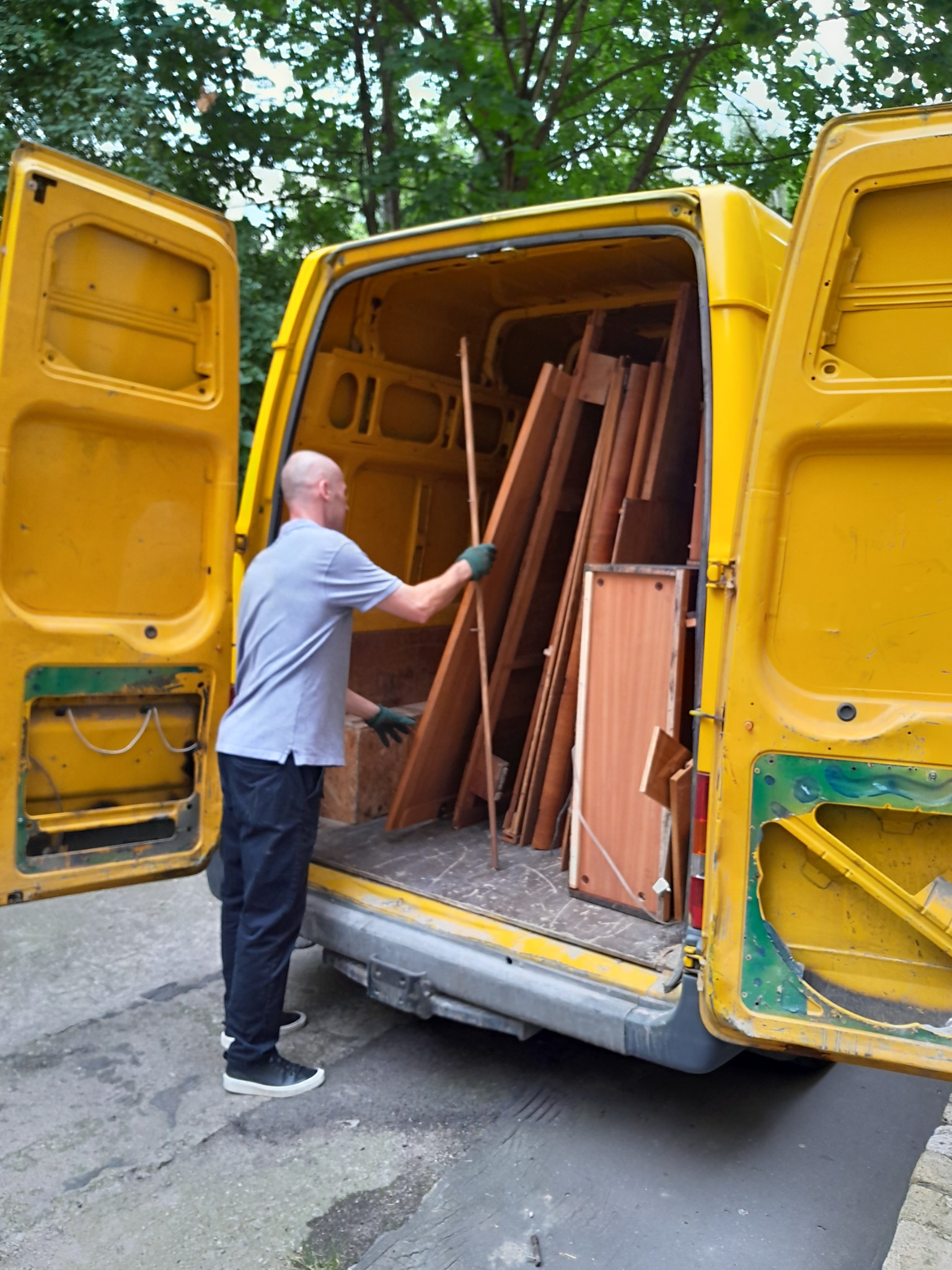 Mężczyzna w rękawicach wkłada drewniane elementy meblowe do otwartego, żółtego busa z widocznymi śladami użytkowania.