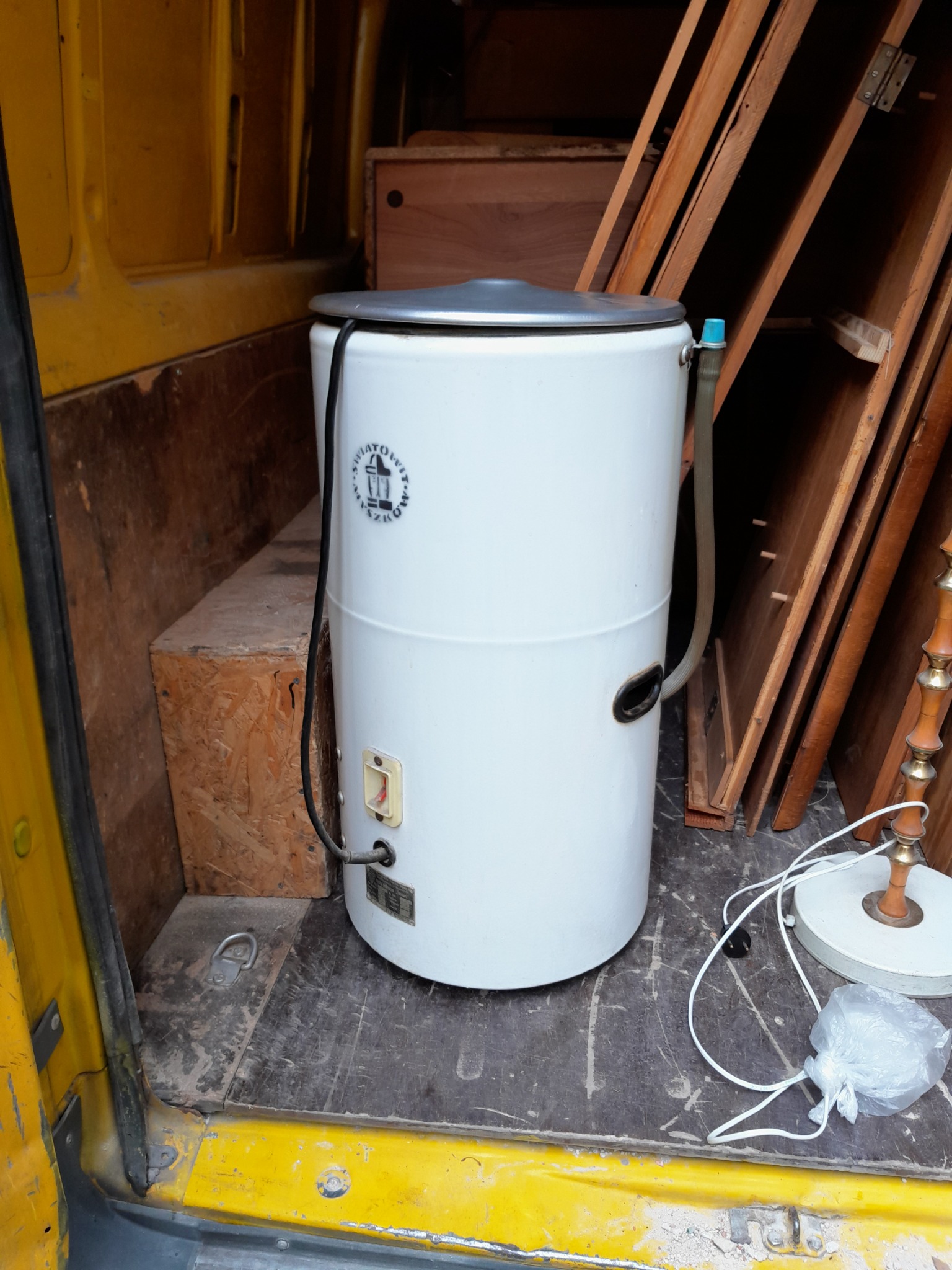 Wnętrze żółtego busa z załadowaną starą, białą pralką wirnikową marki ŚWIATOWIT, obok drewniane elementy i lampa z abażurem przygotowane do przewozu.