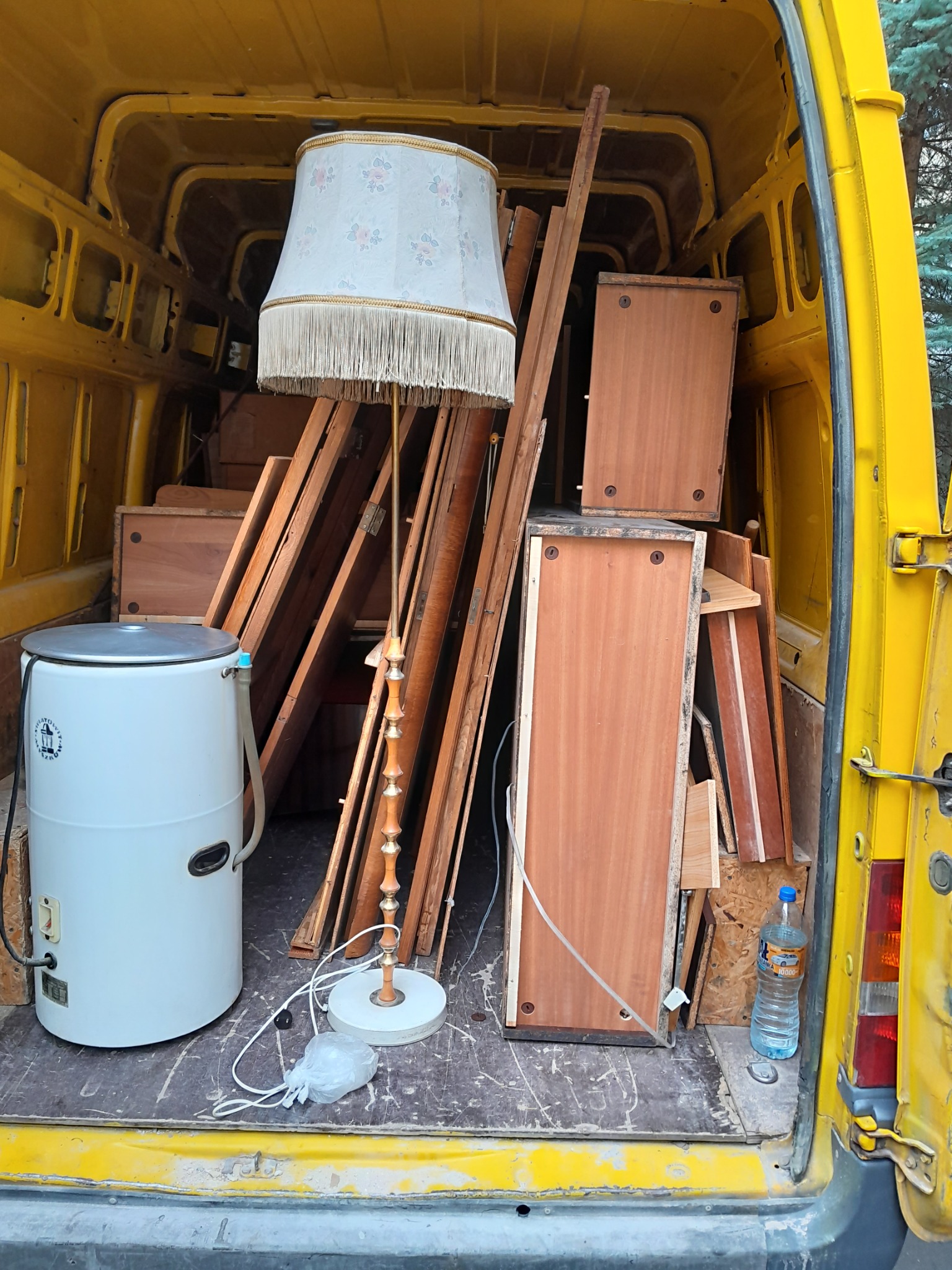 Wnętrze żółtego busa z Wrocławia wypełnione meblami do transportu: szafki, deski, lampa podłogowa z abażurem i stara pralka automatyczna.