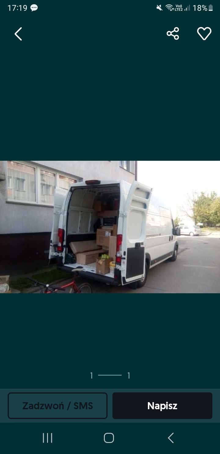 Biały bus z otwartymi drzwiami bagażnika, wypełniony kartonami i przedmiotami, zaparkowany przed budynkiem.