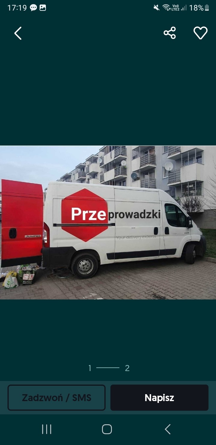 Biały samochód dostawczy z otwartymi czerwonymi drzwiami z tyłu, na których widnieje napis Przeprowadzki w czerwonym rombie, zaparkowany na tle bloku mieszkalnego.