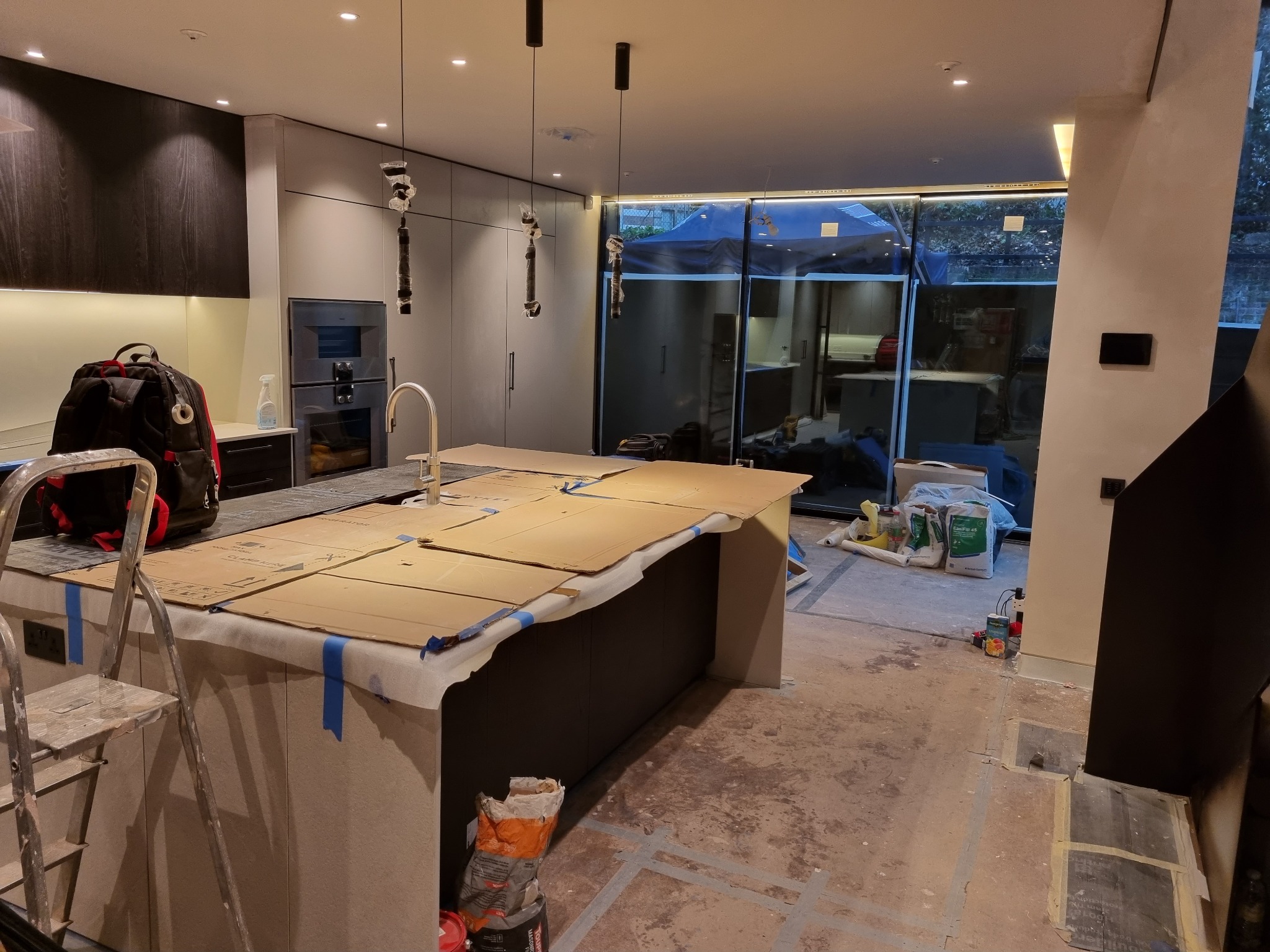 Wnętrze kuchni w trakcie remontu: wyspa kuchenna zabezpieczona kartonem i taśmą, drabina z torbą narzędziową obok, wiszące lampy w folii ochronnej, widoczne puszki instalacyjne na ścianie, worki...