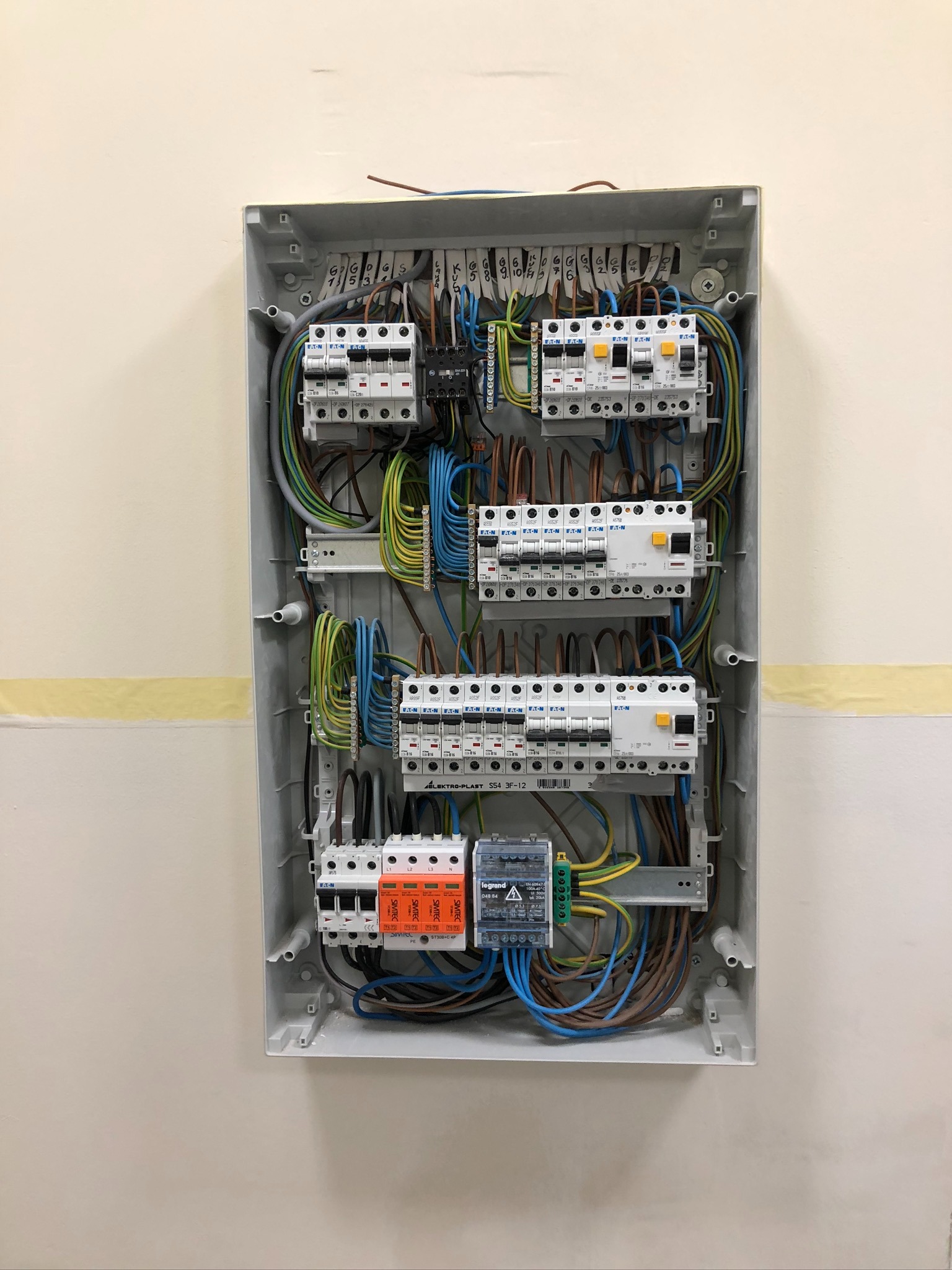 Otwarta skrzynka elektryczna z bezpiecznikami, przewodami oznaczonymi numerami i różnymi elementami instalacji, zamontowana na ścianie z taśmą malarską. Widoczne bezpieczniki Eaton i Legrand.