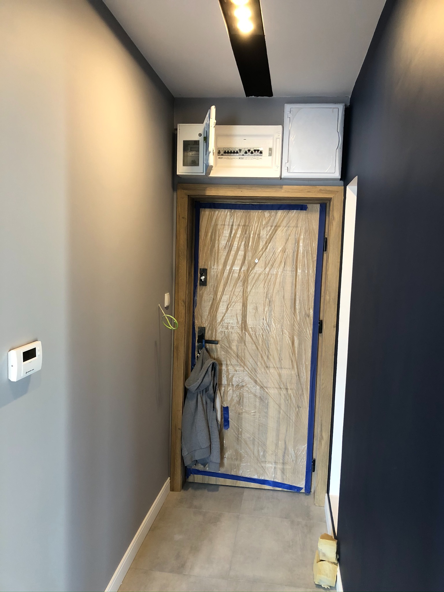 Korytarz w trakcie remontu: zabezpieczone folią drzwi, otwarta skrzynka z bezpiecznikami nad nimi, szare ściany, widoczny termostat i fragment garderoby na wieszaku.