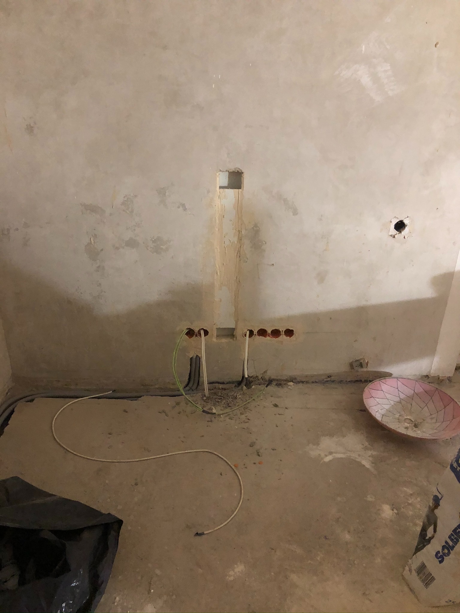 Surowa ściana z widocznymi przewodami elektrycznymi wystającymi z otworów, przygotowana do dalszych prac wykończeniowych, widoczny worek z zaprawą na podłodze.