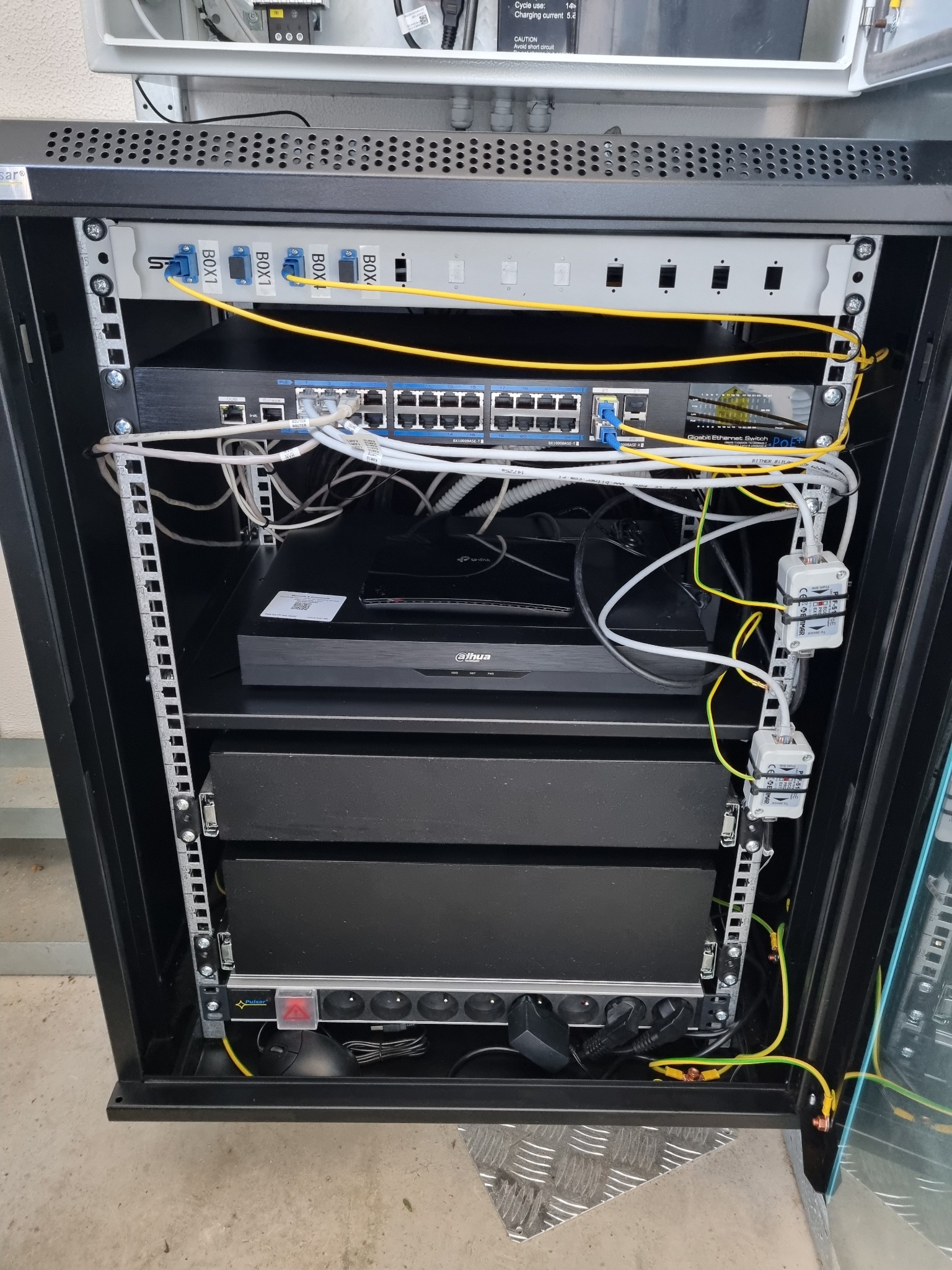 Szafa rack z zamontowanymi urządzeniami sieciowymi: patch panel światłowodowy, switch Gigabit Ethernet PoE, rejestrator cyfrowy Dahua i listwa zasilająca Pulsar, okablowanie strukturalne.