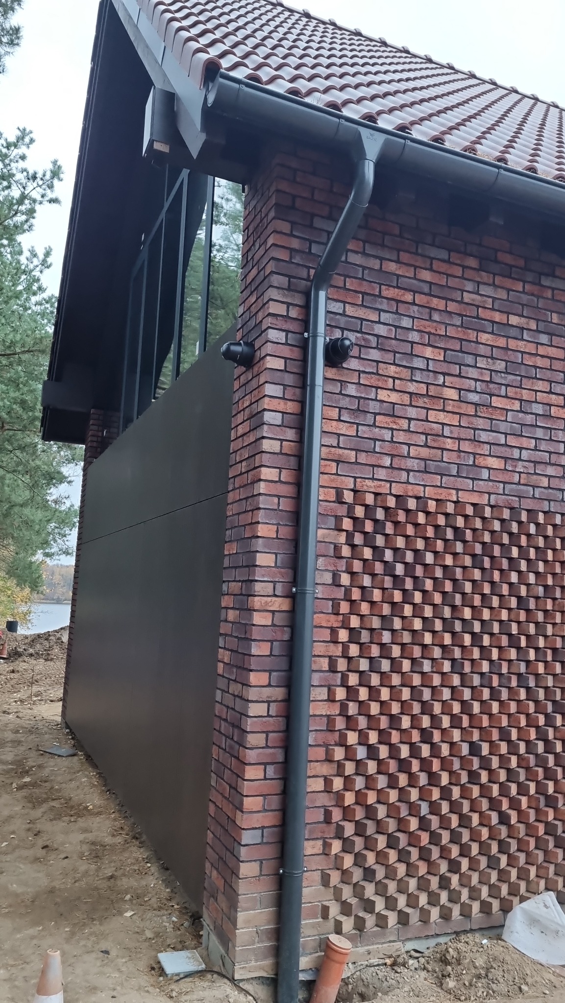 Zewnętrzny narożnik budynku z czerwonej cegły z dekoracyjnym wzorem, widoczna rynna spustowa i dwa czujniki, prawdopodobnie kamery, na tle jeziora i drzew