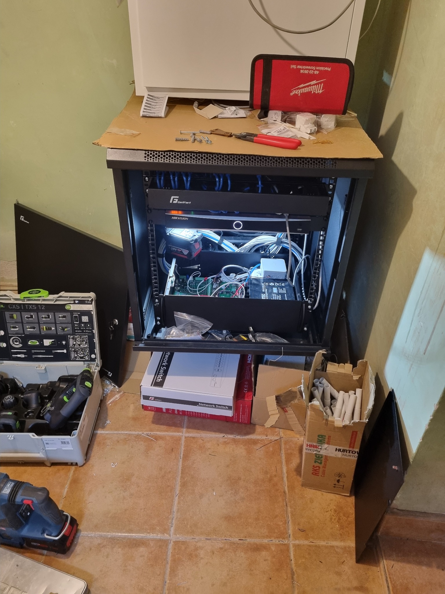 Szafa rack z okablowaniem sieciowym, narzędzia Festool i Milwaukee. Widoczne switche GetFort i Hikvision. Instalacja w trakcie realizacji, na podłodze płytki.