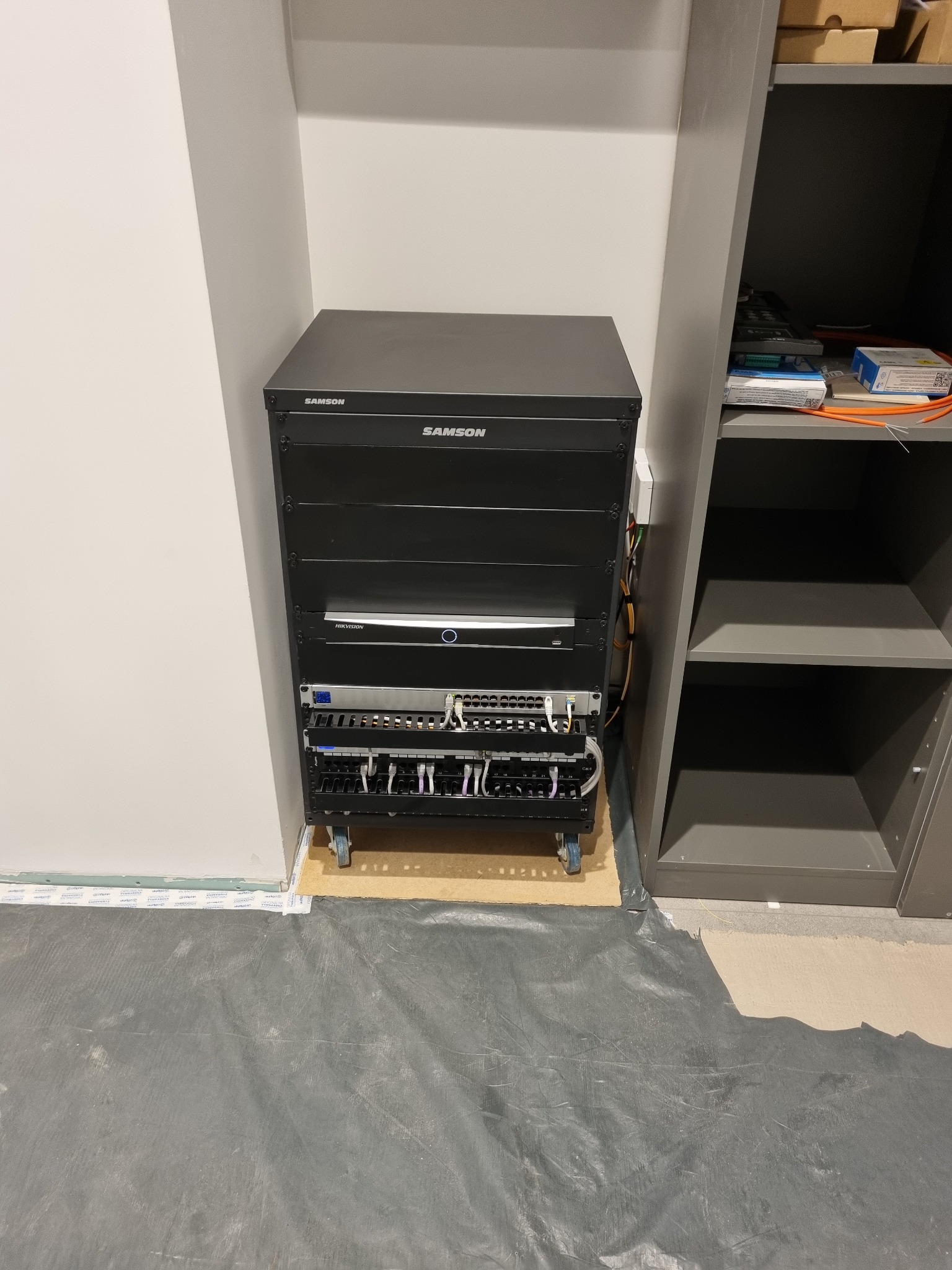 Czarna szafa rack firmy Samson z zamontowanym sprzętem sieciowym (router, switche) w pomieszczeniu z szarą podłogą i regałem. Widoczne kable i złącza.