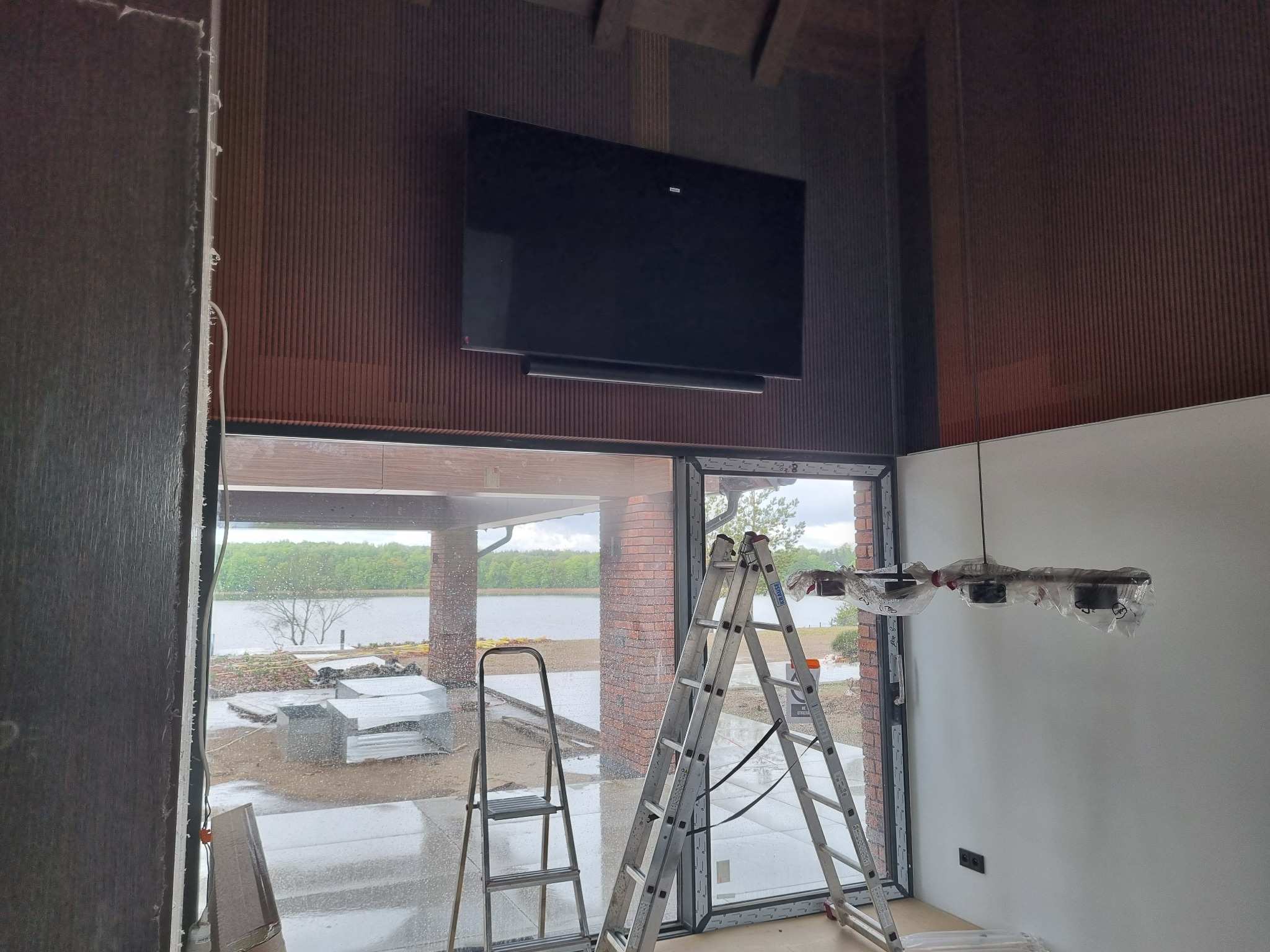 Montaż telewizora na ścianie z dekoracyjnymi panelami. Widoczne dwie drabiny, okno z widokiem na jezioro oraz lampa w folii ochronnej. Prace wykończeniowe w nowym domu.