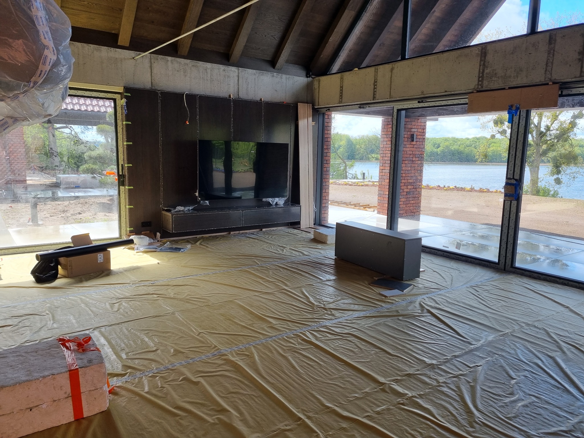 Wnętrze domu w trakcie instalacji elektrycznej, z widocznymi przewodami na ścianie i zabezpieczeniem podłogi folią. Duże okna z widokiem na jezioro.