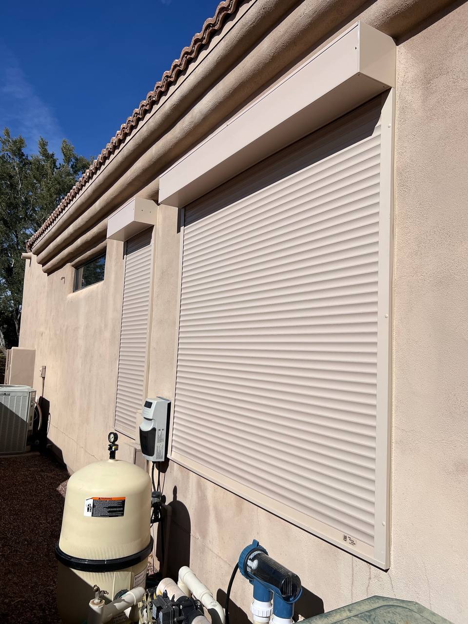 Zamknięte rolety zewnętrzne w kolorze beżowym na elewacji domu z widoczną instalacją basenową i błękitnym niebem w tle.